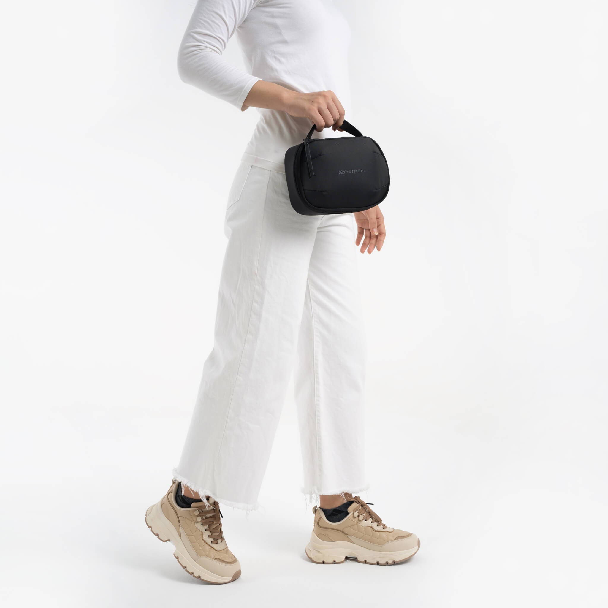 Lower body view of a model dressed in all white holding Sherpani travel accessory the Atlas in Carbon. 