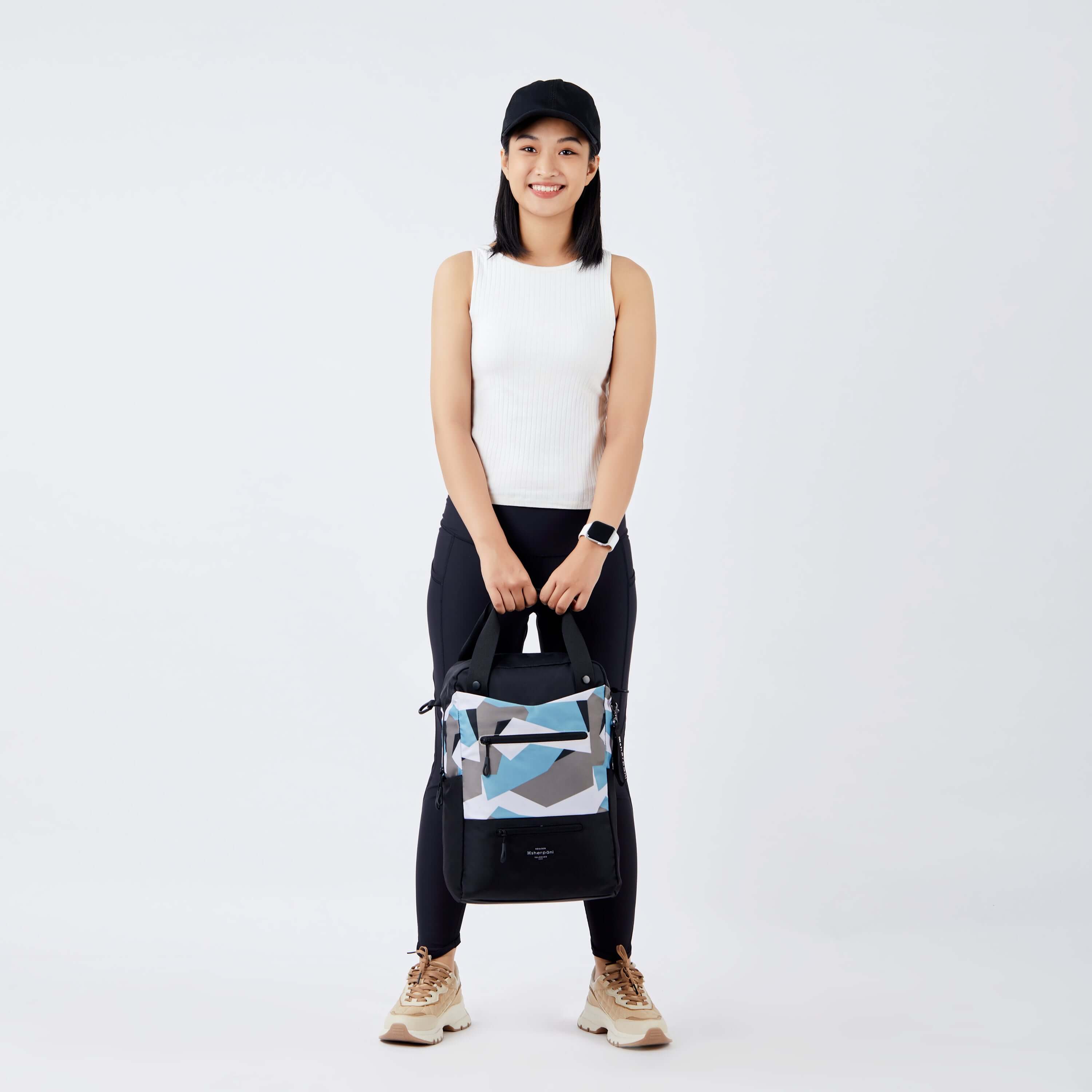 Full body view of a dark haired model smiling at the camera. She is wearing a black hat, white tank top, black leggings and sneakers. She is carrying Sherpani's three in one bag, the Camden in Summer Camo, by the tote handles. 