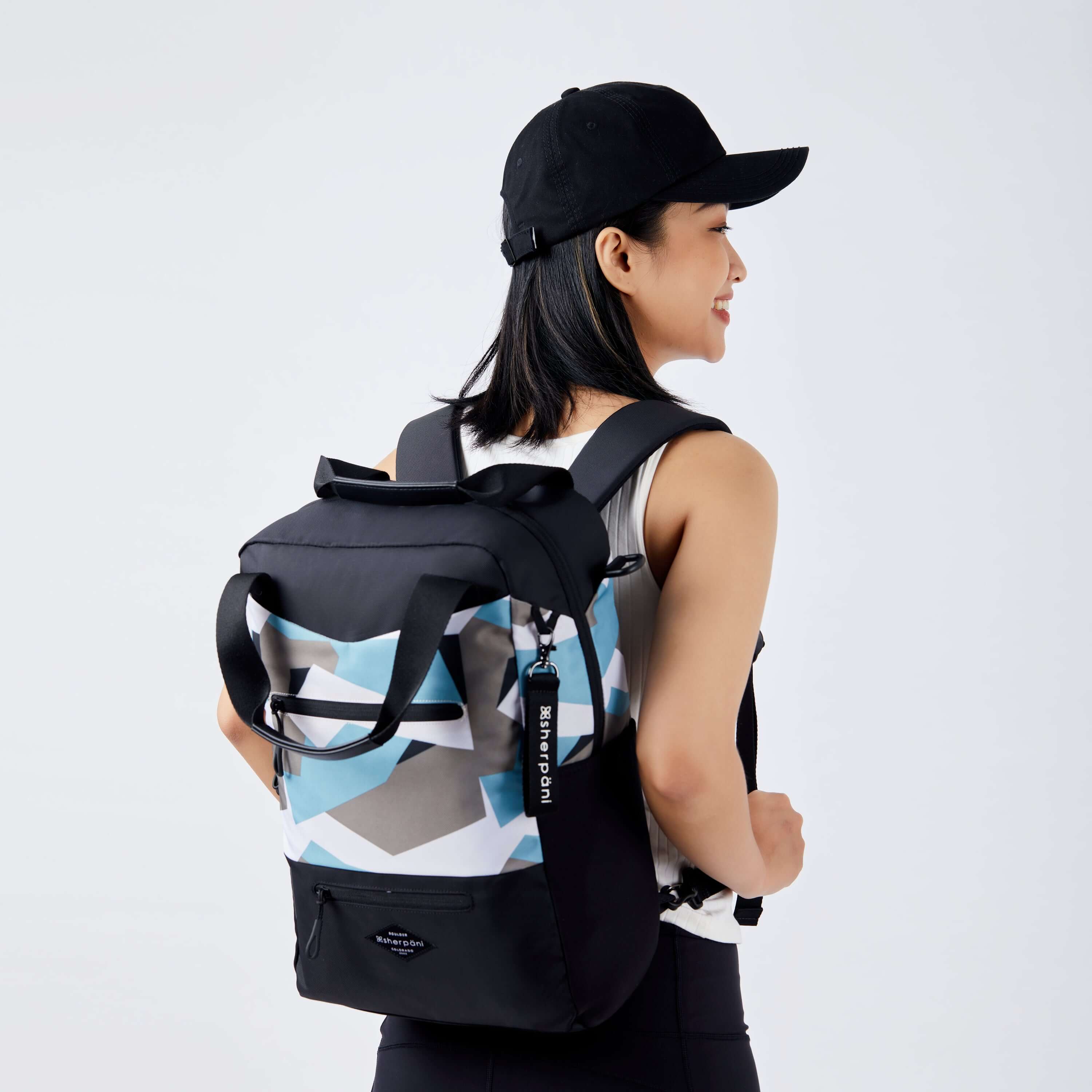 Close up view of a dark haired model facing away from the camera and smiling. She is wearing a black hat, white tank top and black leggings. She is carrying Sherpani's three in one bag, the Camden in Summer Camo, as a backpack. #color_summer camo