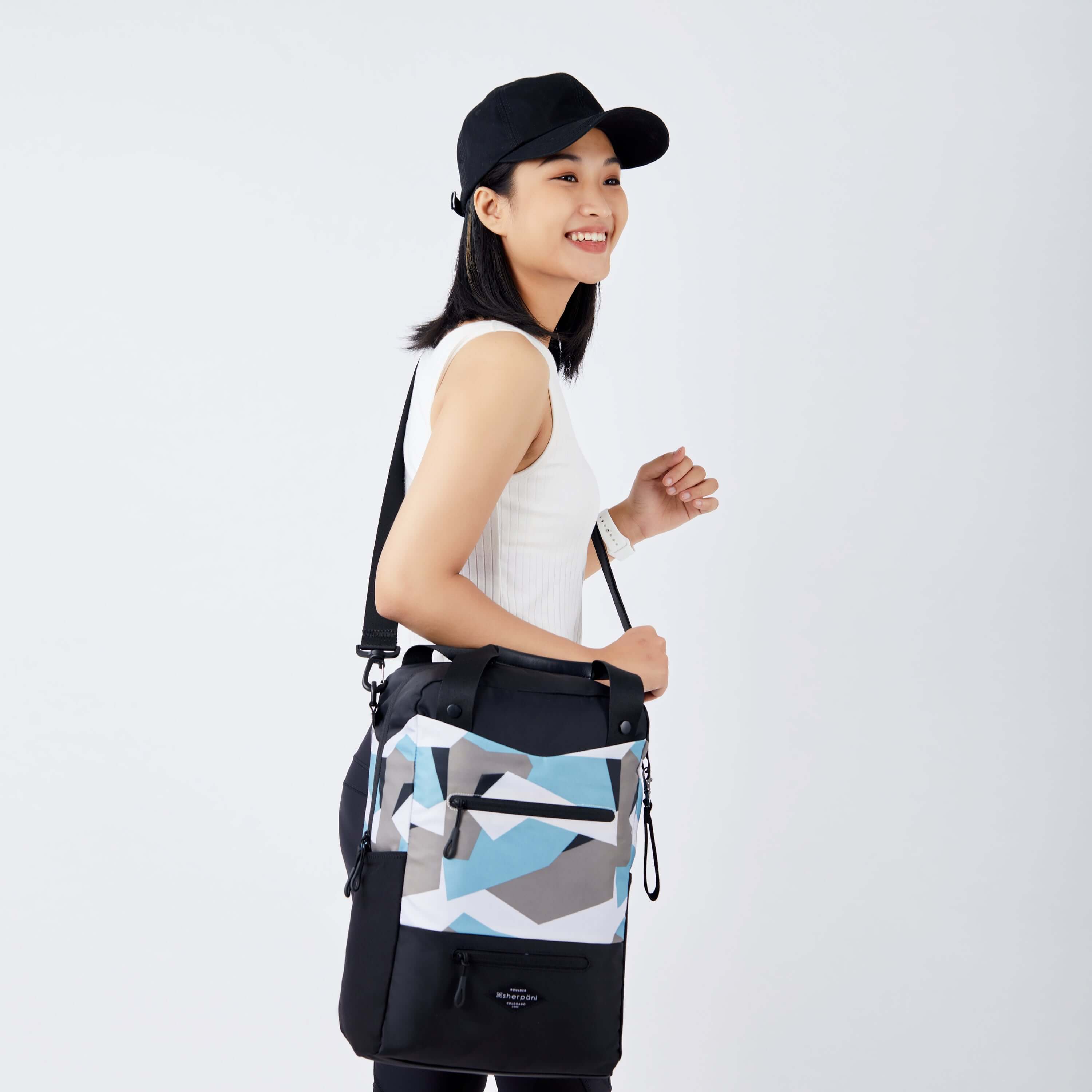 Close up view of a dark haired model facing to the side and smiling. She is wearing a black hat, white tank top and black leggings. She is carrying Sherpani's three in one bag, the Camden in Summer Camo, as a crossbody. 
