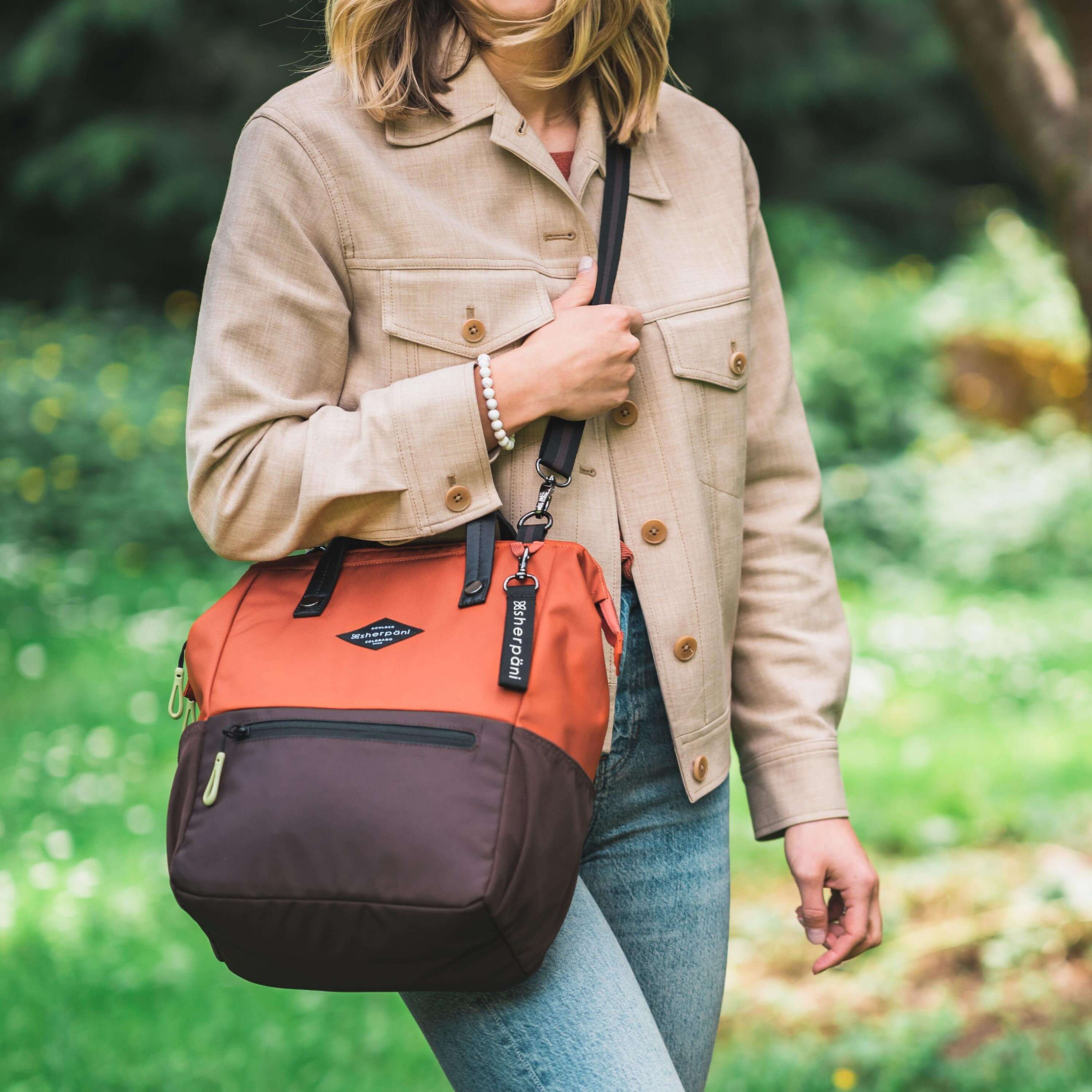 A blonde woman stands outside in a park. She is wearing a tan jacket and jeans. She carries Sherpani three-in-one bag, the Dispatch in Clay, as a crossbody. #color_clay