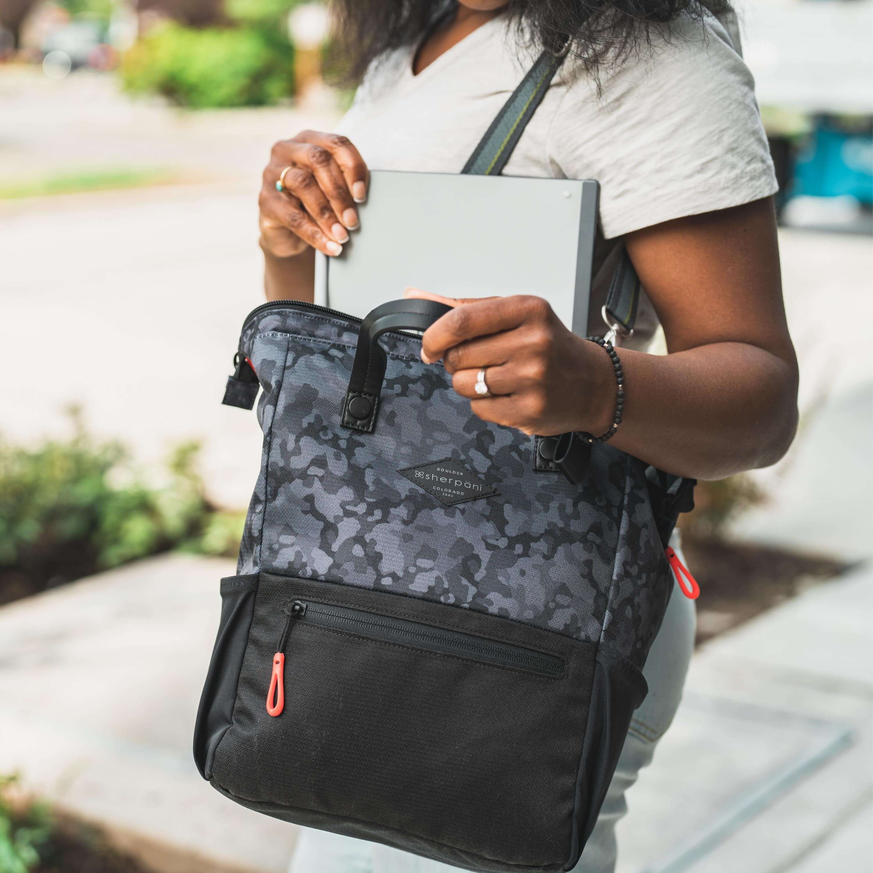 Close up view of a curly haired woman standing on a street. She is pulling her laptop out of Sherpani three in one bag, the Dispatch in Dream Camo, which she carries as a crossbody. 