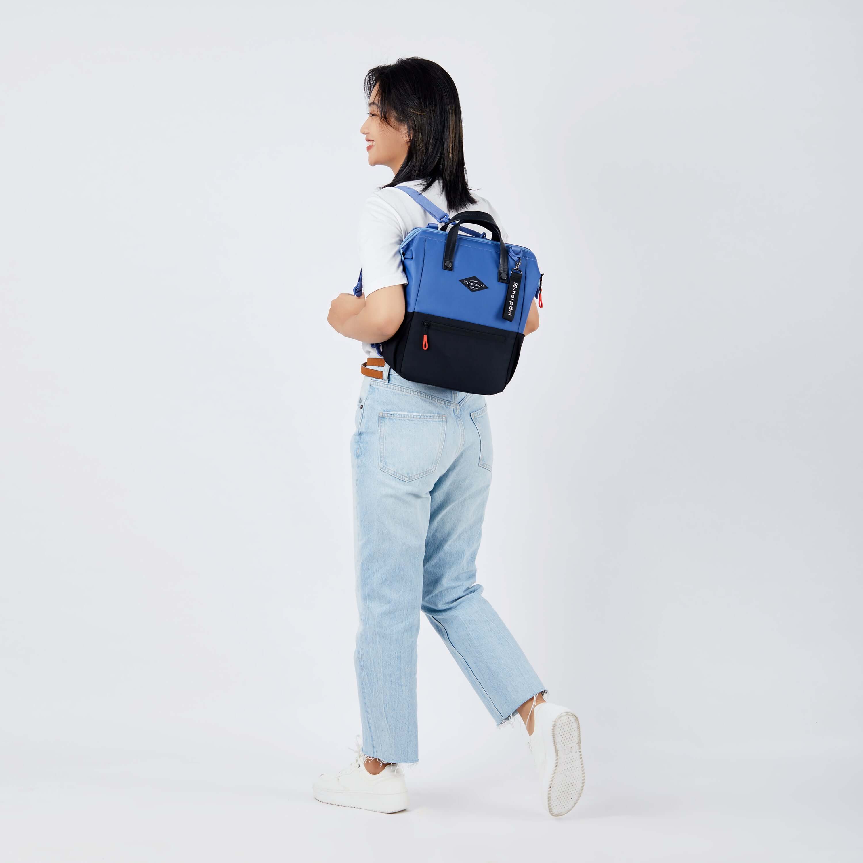 Full body view of a dark haired model facing away from the camera and walking. She is wearing a white tee shirt, jeans and white sneakers. She carries Sherpani three in one bag, the Dispatch in Pacific Blue, as a backpack. 