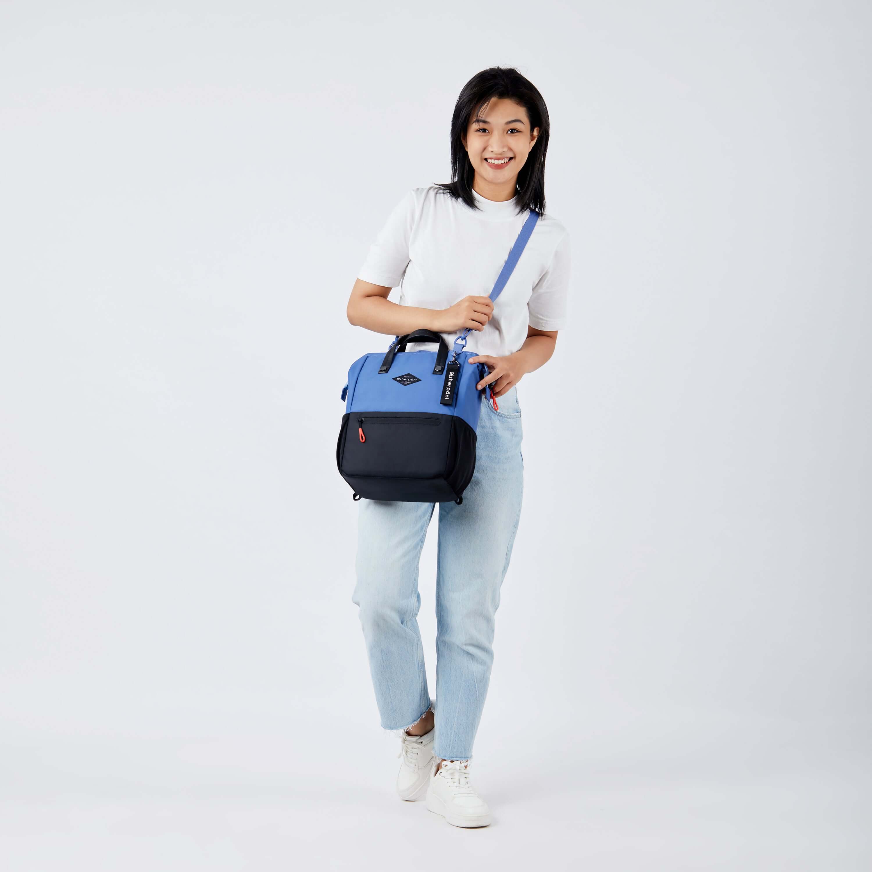 Full body view of a dark haired model facing the camera and smiling. She is wearing a white tee shirt, jeans and white sneakers. She carries Sherpani three in one bag, the Dispatch in Pacific Blue, as a crossbody. 