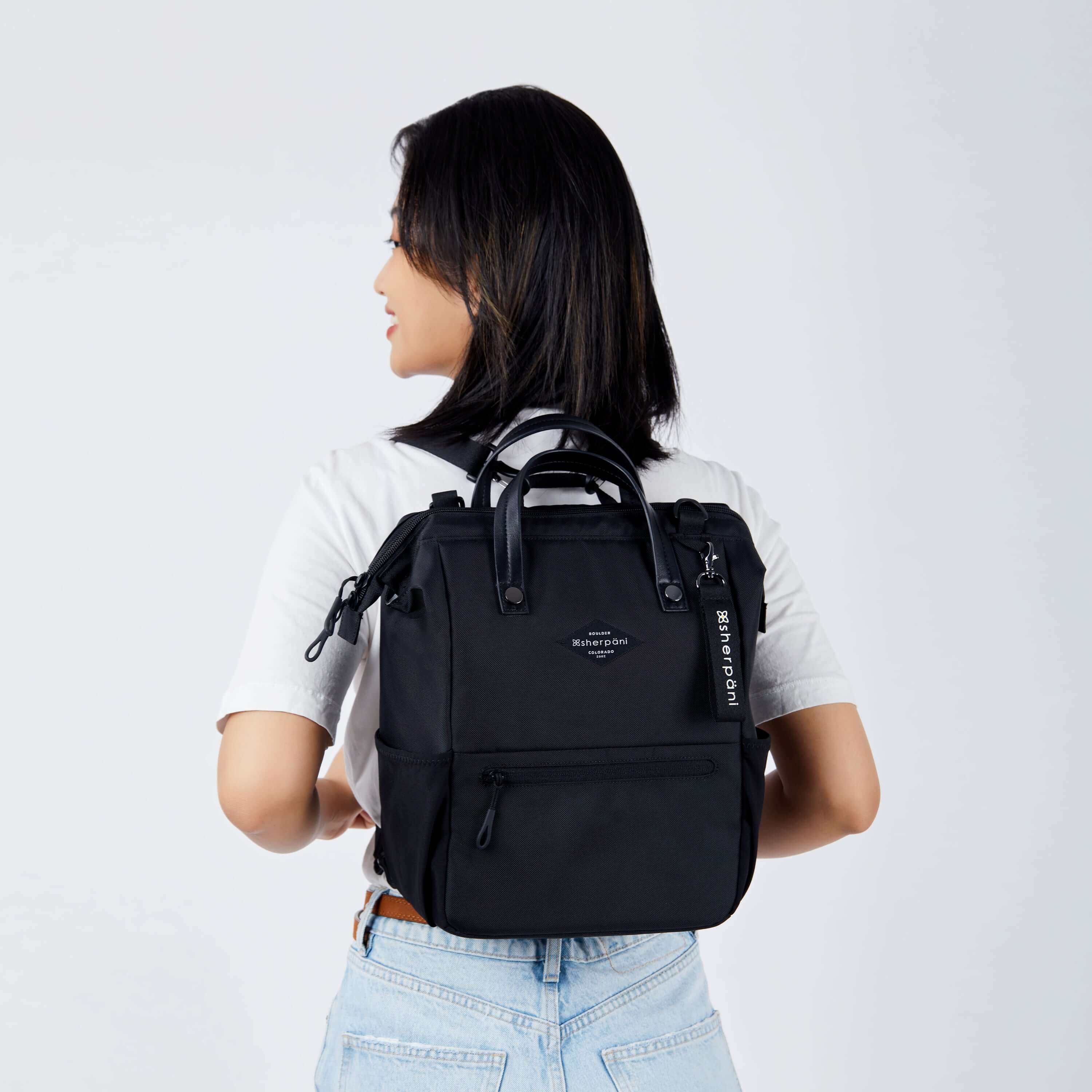 Close up view of a dark haired model facing away from the camera and looking over her left shoulder. She is wearing a white tee shirt and jeans. She carries Sherpani three in one bag, the Dispatch in Raven, as a backpack. 