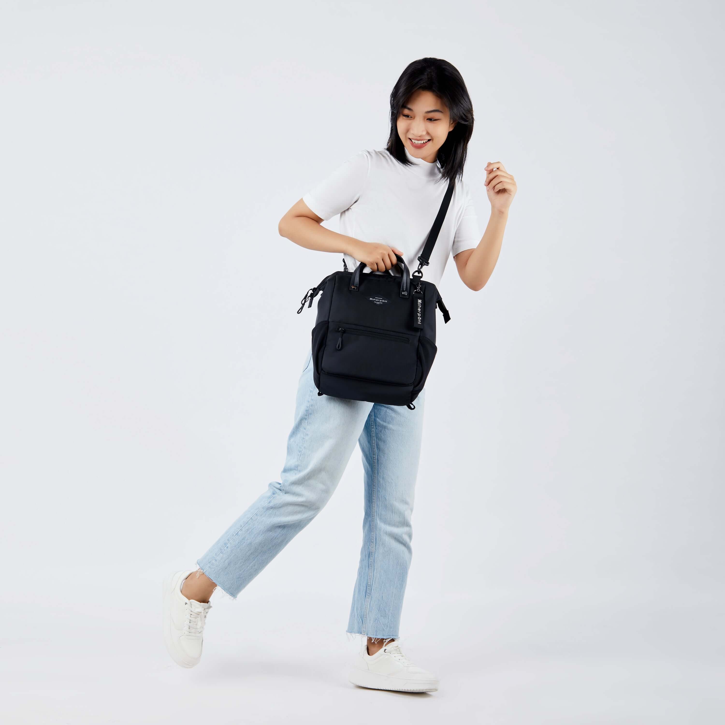 Full body view of a dark haired model facing the side and smiling downward. She is wearing a white tee shirt, jeans and white sneakers. She carries Sherpani three in one bag, the Dispatch in Raven, as a crossbody. 