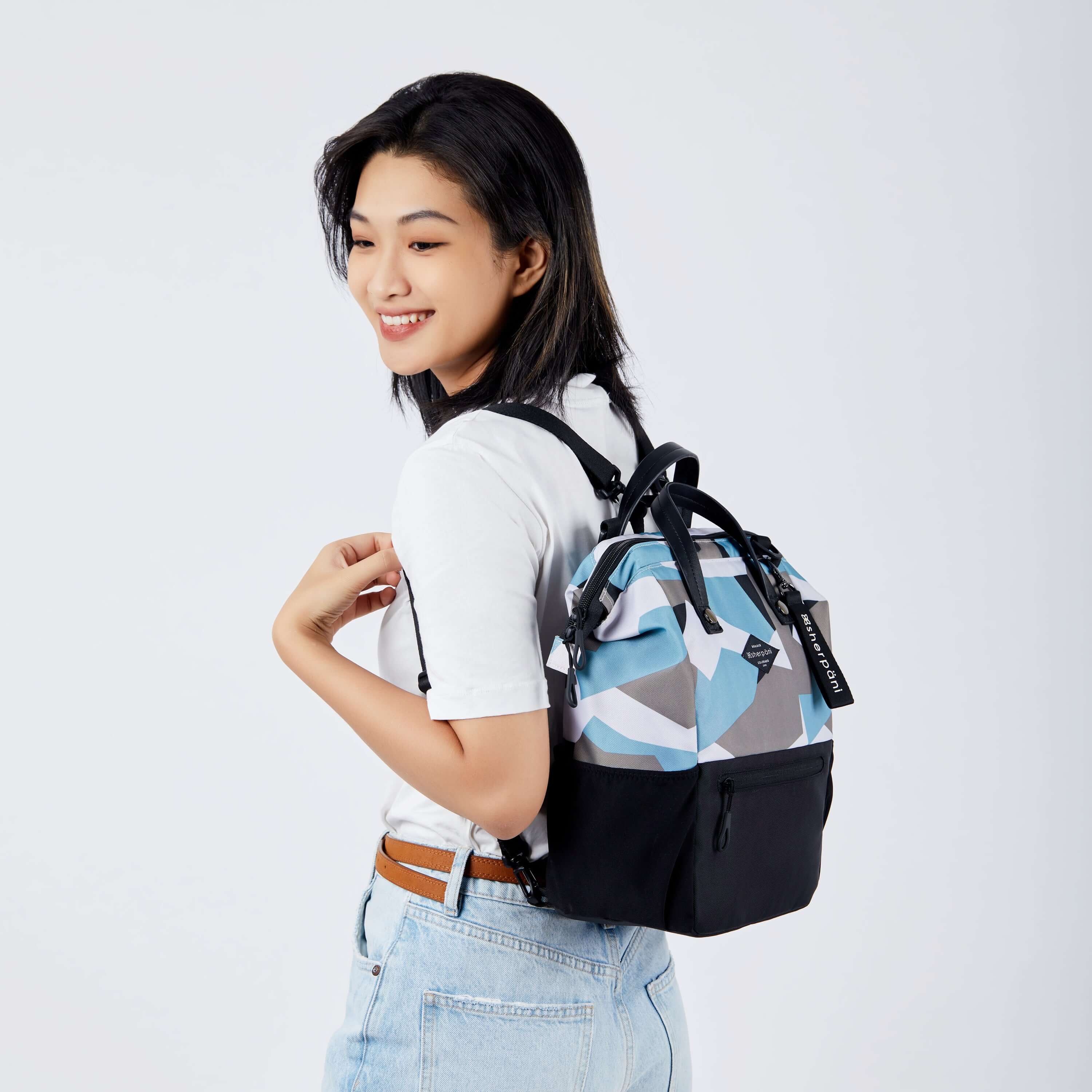 Close up view of a dark haired model facing away from the camera and smiling over her left shoulder. She is wearing a white tee shirt and jeans. She carries Sherpani three in one bag, the Dispatch in Summer Camo, as a backpack. 