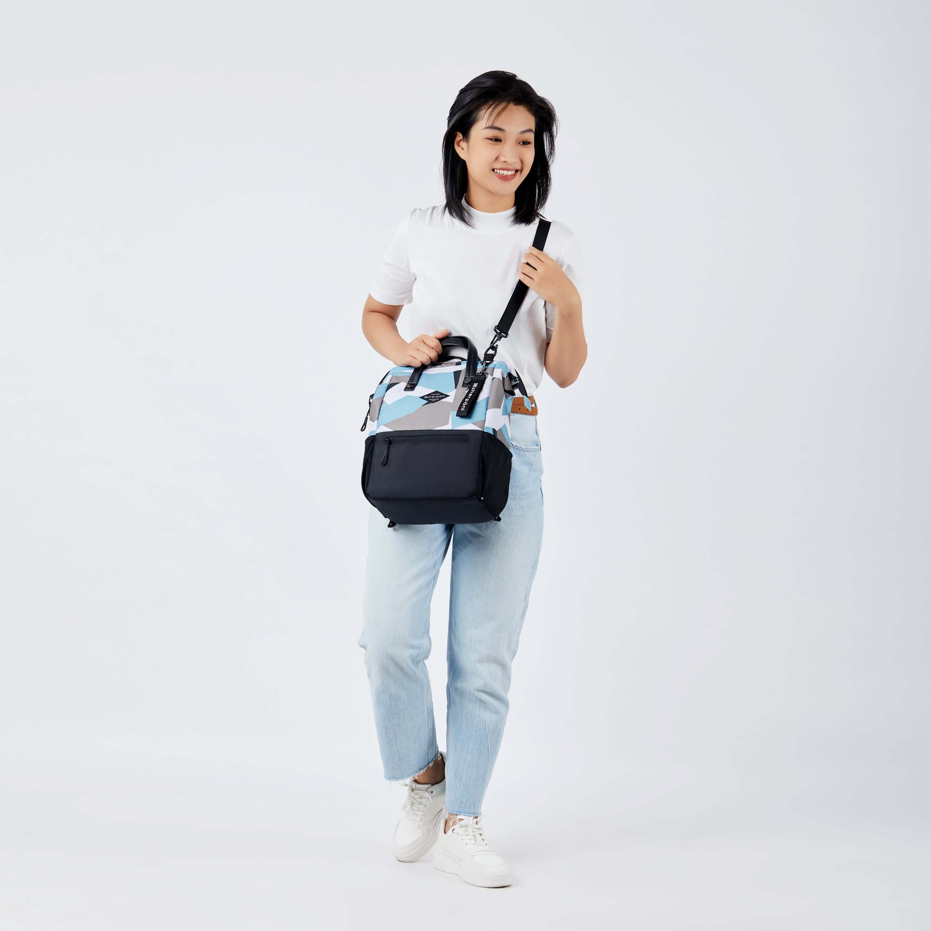 Full body view of a dark hair model facing the camera and smiling to the side. She is wearing a white tee shirt, jeans and white sneakers. She carries Sherpani three in one bag, the Dispatch in Summer Camo, as a crossbody. 