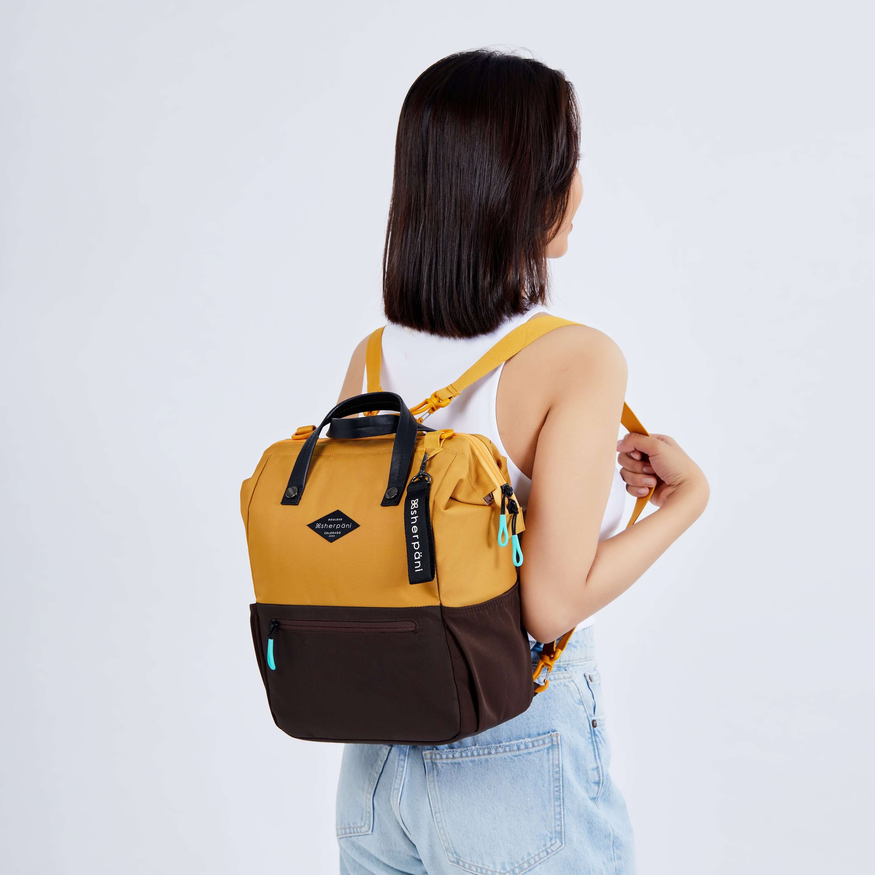 Close up view of a dark haired model facing away from the camera. She is wearing a white tank top and jeans. She carries Sherpani three in one bag, the Dispatch in Sundial, as a backpack. 