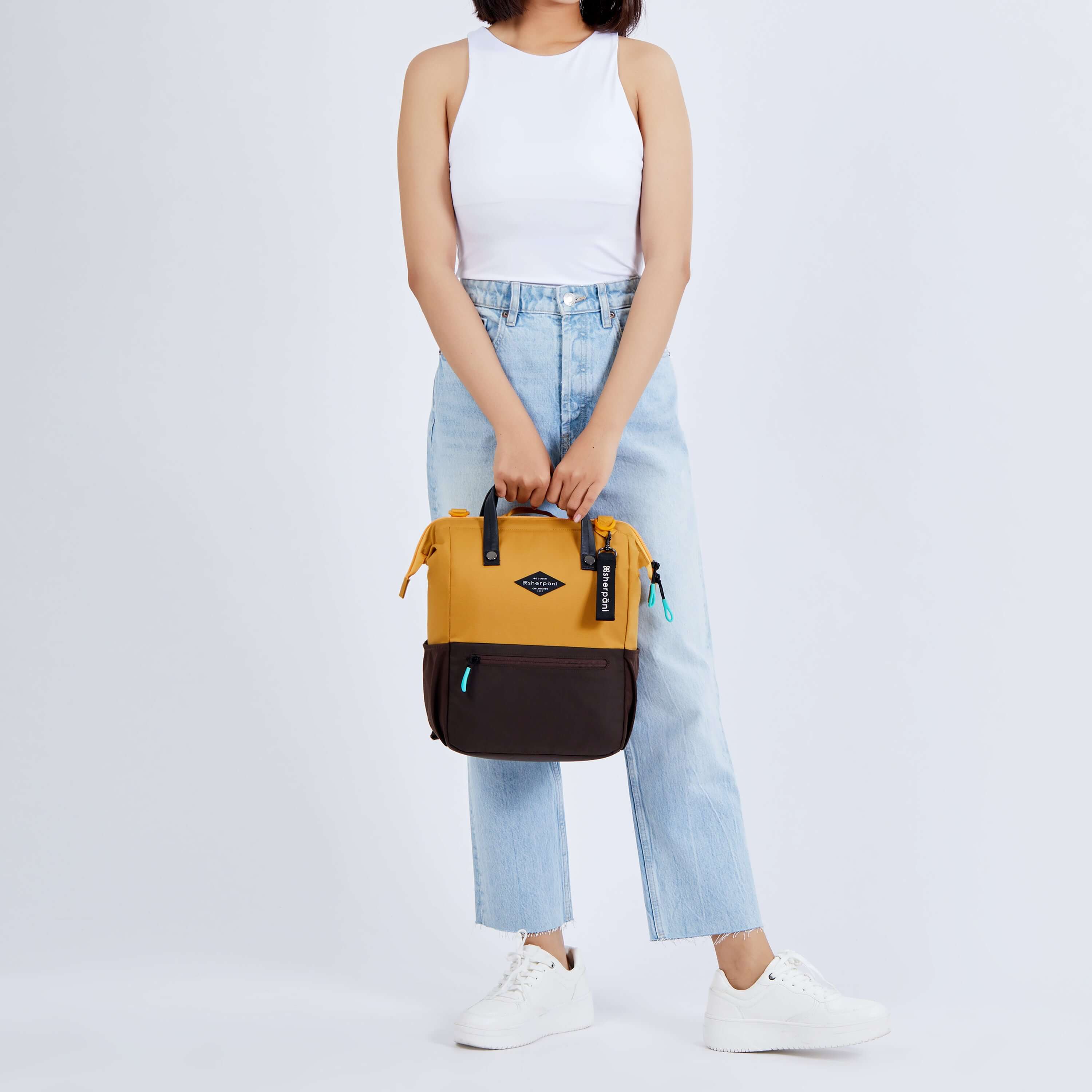 Full body view of a dark haired model facing the camera. She is wearing a white tank top, jeans and white sneakers. She carries Sherpani three in one bag, the Dispatch in Sundial, by its tote handles. 