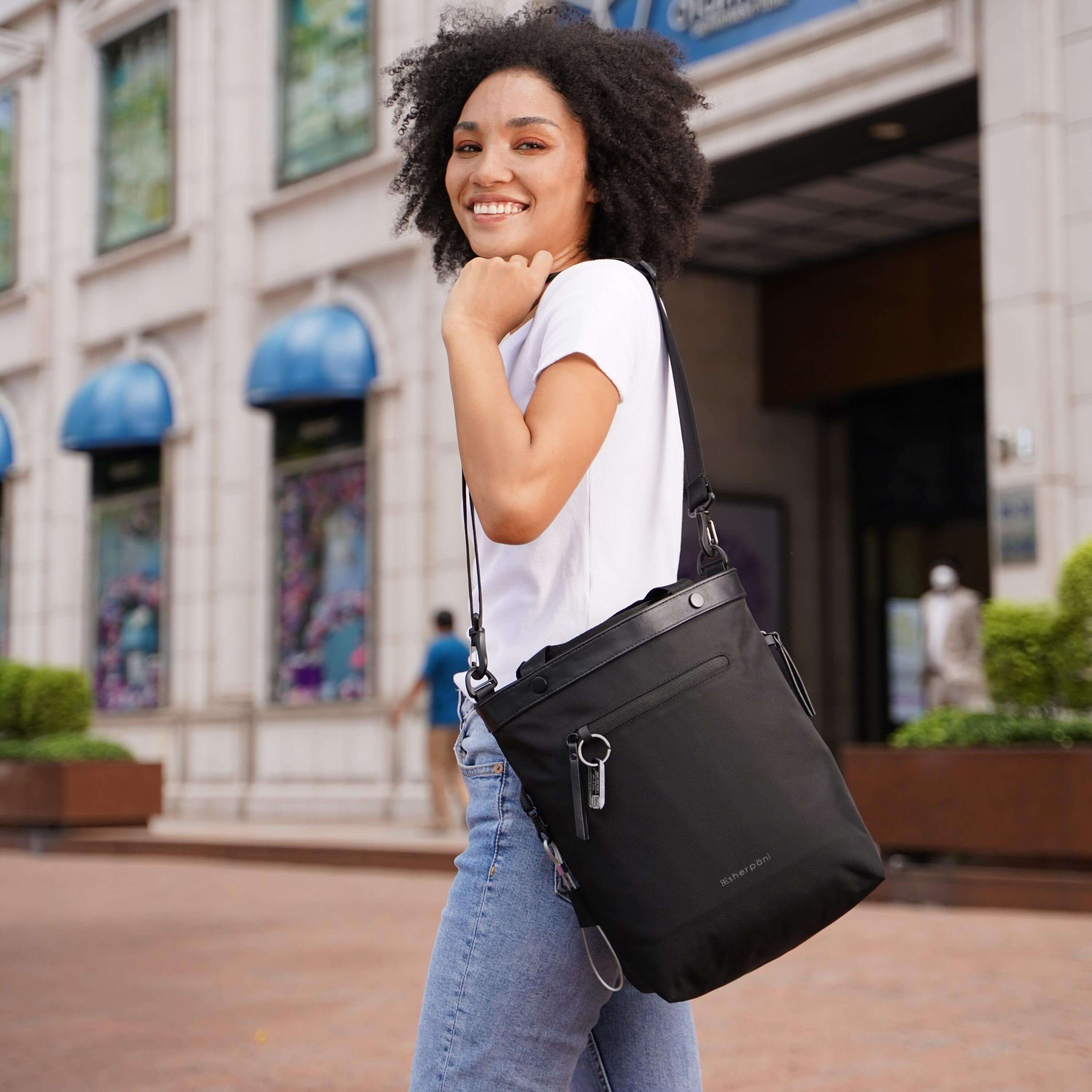 A curly haired model is standing on the street of an outdoor shopping mall. She is wearing a white tee shirt and jeans, and is smiling at the camera. Over her shoulder is Sherpani's Anti-Theft bag, the Geo AT in Carbon. 