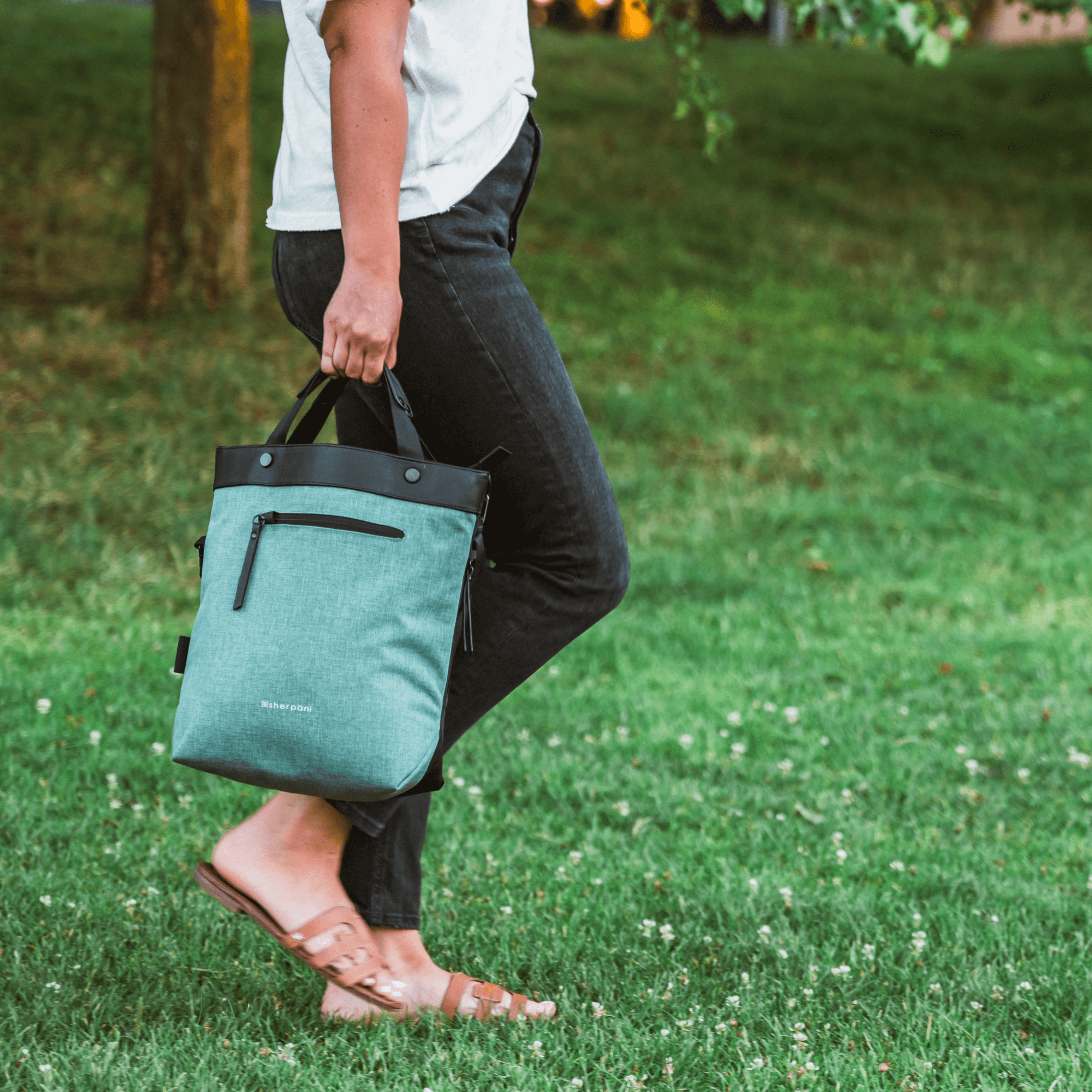 Close up view of a model's lower body. She is facing the side and walking on the grass. She wears a white shirt, black pants and tan sandals. She is carrying Sherpani's Ant-Theft bag, the Geo AT in Teal, by the tote handles. 