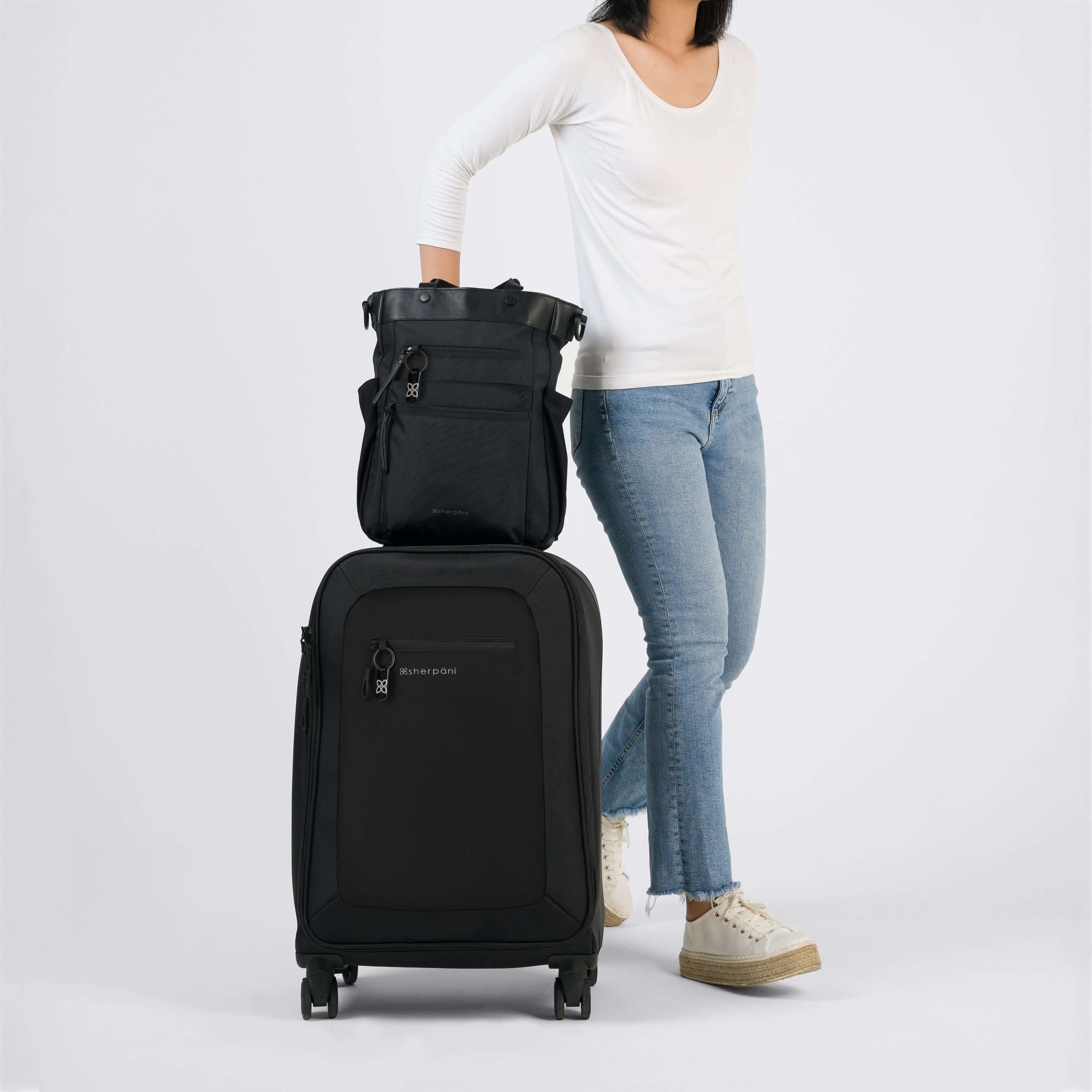 Lower body view of a dark haired model facing the camera. She is wearing a white shirt, jeans and white shoes. Next to her stands Sherpani's Anti-Theft luggage, the Hemisphere in Carbon. On top of the suitcase sits Sherpani's Anti-Theft bag, the Soleil in Carbon, utilizing the luggage-pass through. 