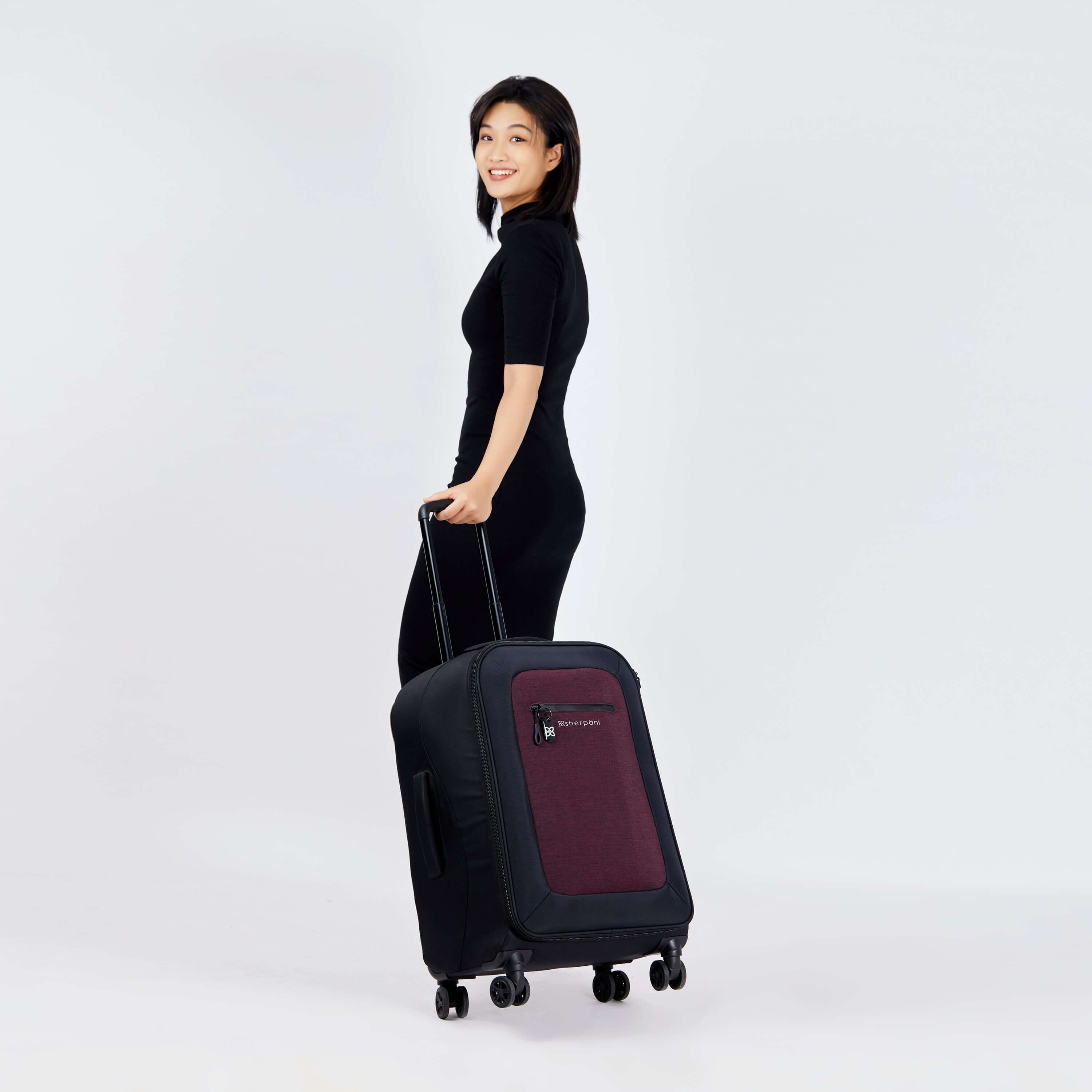 Full body view of a dark haired model smiling over her left shoulder at the camera. She is wearing a black dress. She pulls Sherpani's Anti-Theft luggage, the Hemisphere in Merlot, along beside her. 