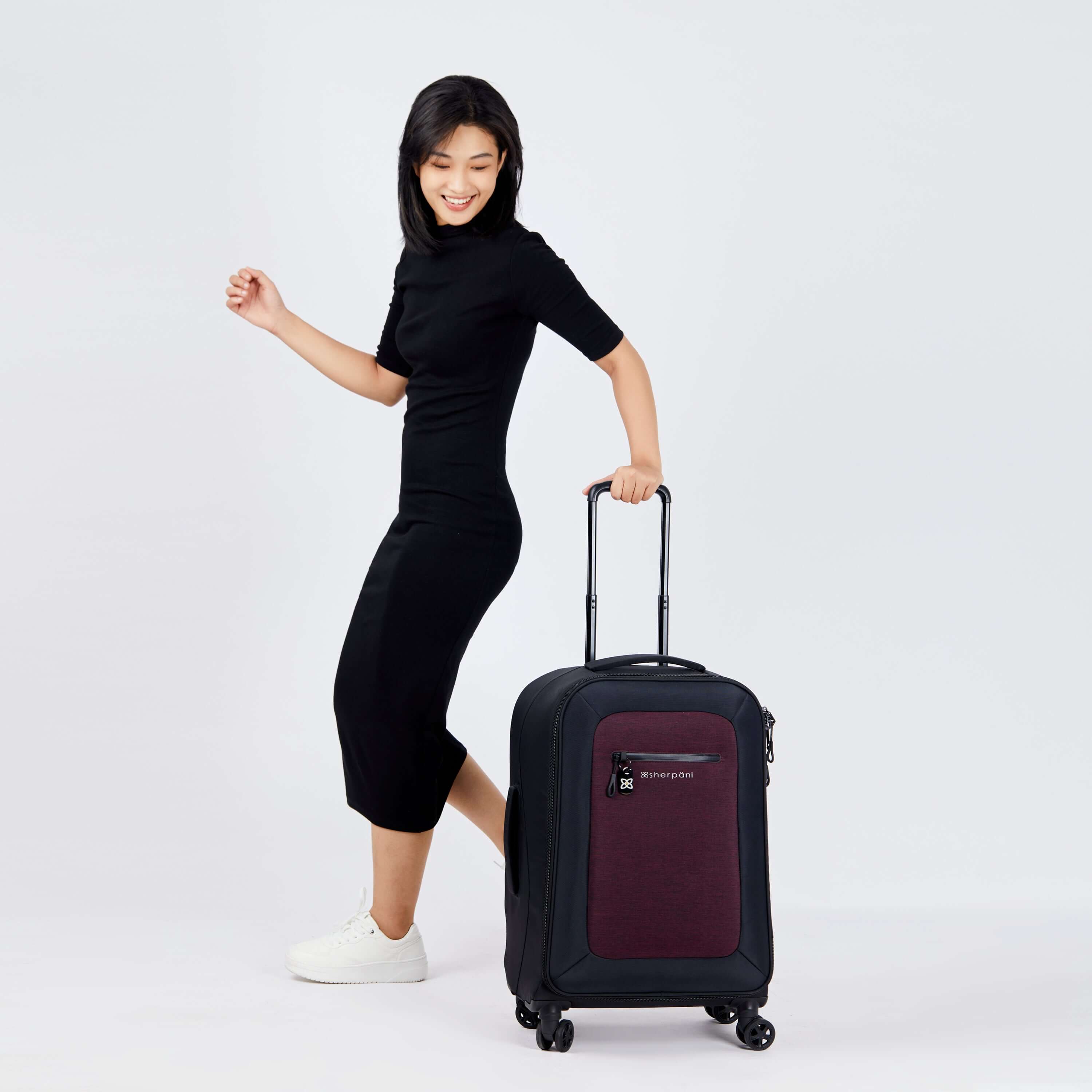 Full body view of a dark haired model facing to the side and smiling downward. She is wearing a black dress and white sneakers. Her left hand grabs the handle of Sherpani's Anti-Theft luggage, the Hemisphere in Merlot, which stands beside her. 