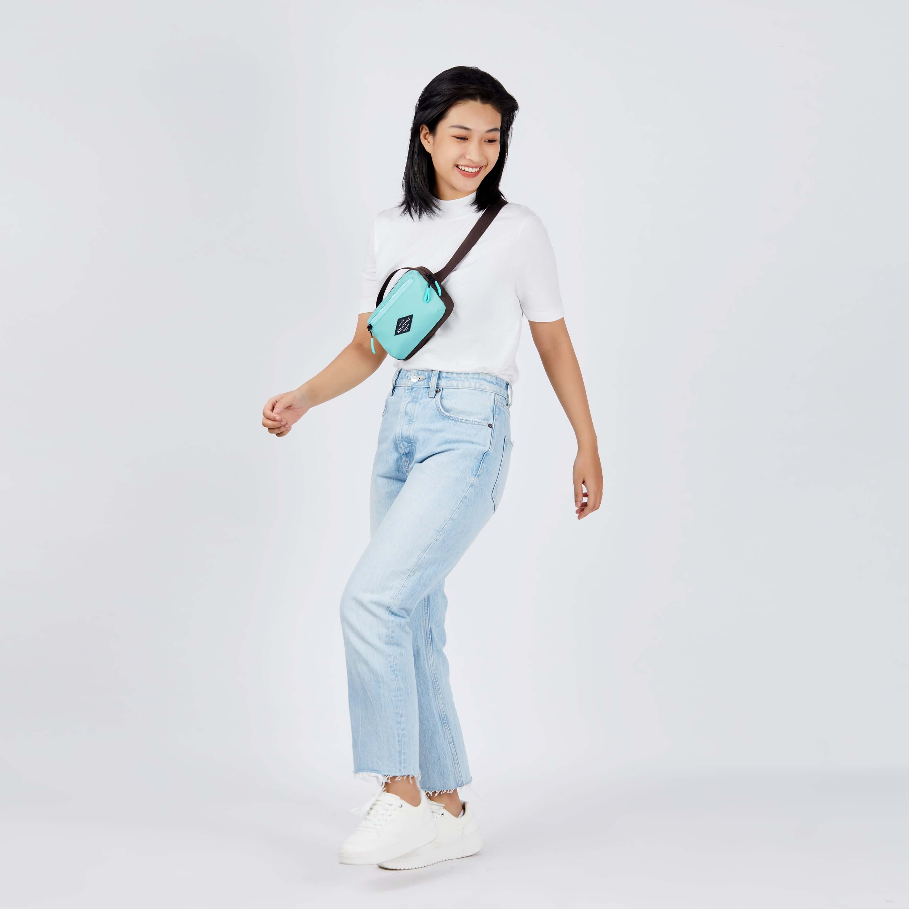 Full body view of a dark haired model smiling over her left shoulder. She is wearing a white tee shirt, faded jeans, white shoes and Sherpani's fanny pack, the Hyk in Seagreen, as a crossbody. 
