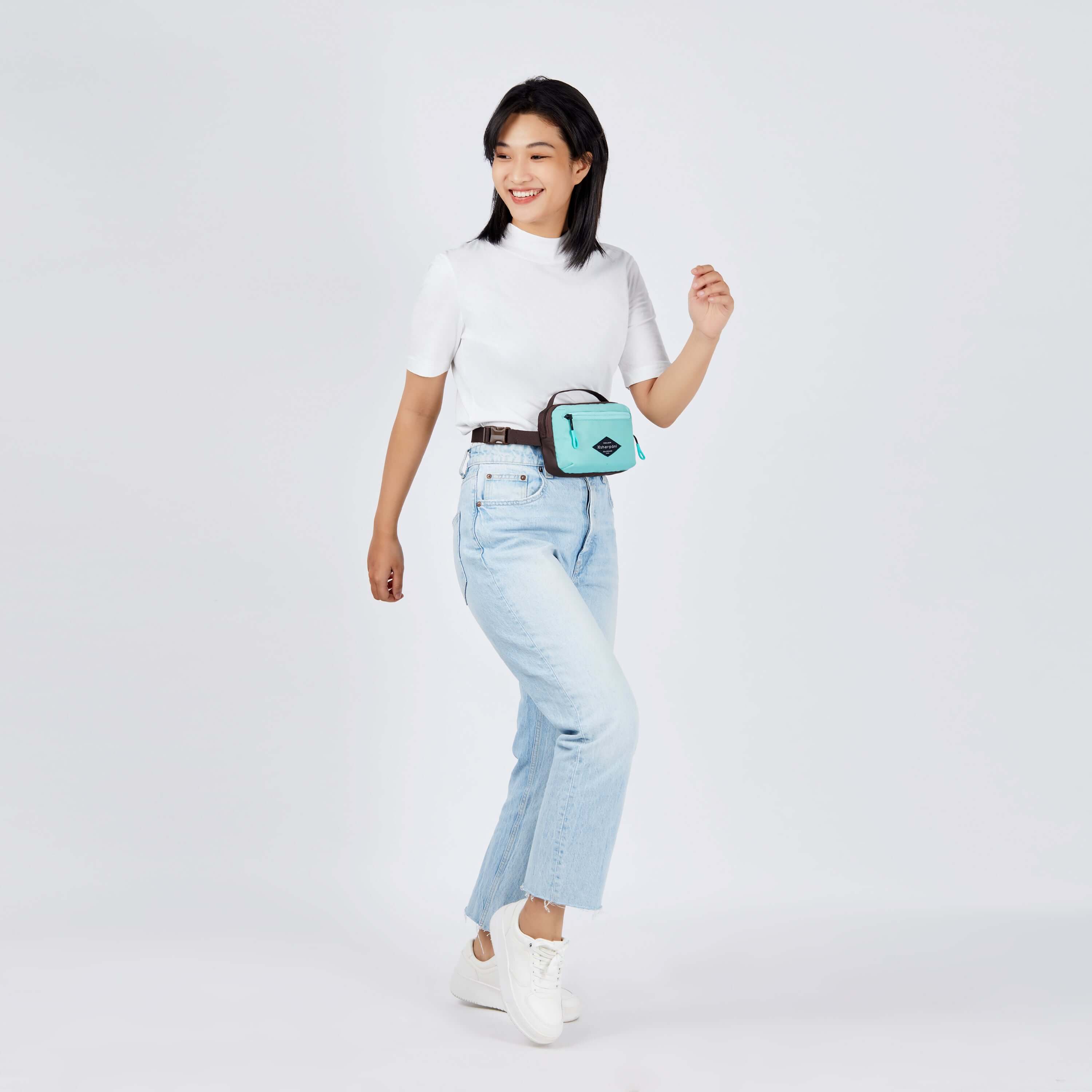 Full body view of a dark haired model smiling over her right shoulder. She is wearing a white tee shirt, faded jeans, white shoes and Sherpani's fanny pack, the Hyk in Seagreen, as a fanny pack. #color_seagreen