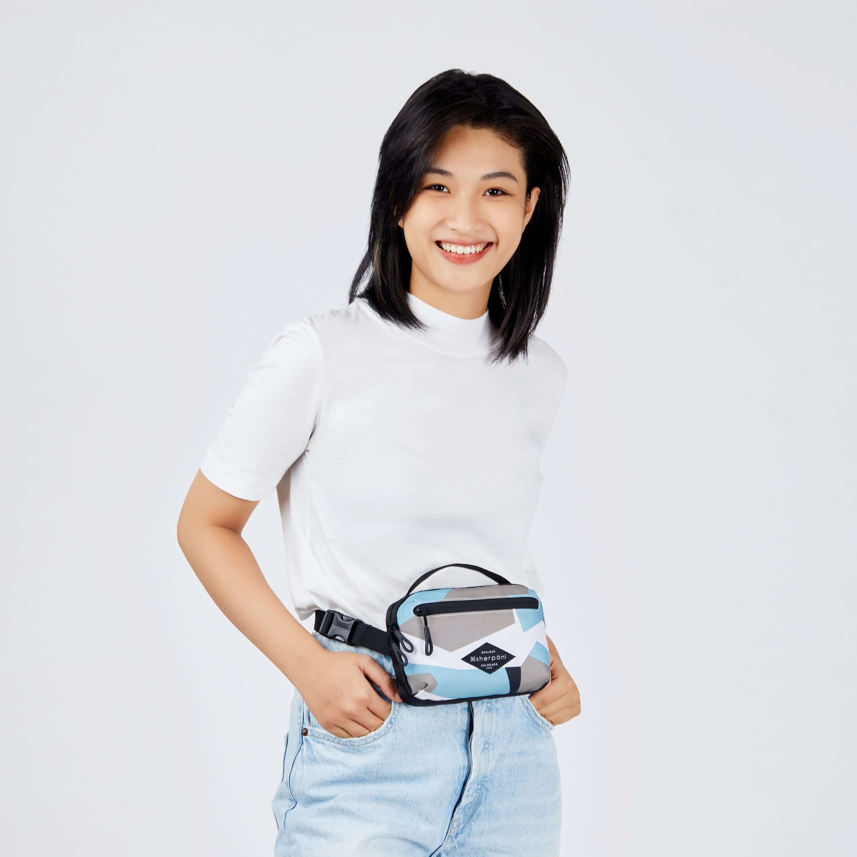 Close up view of a dark haired model facing the camera and smiling. She is wearing a white tee shirt, faded jeans and Sherpani's fanny pack, the Hyk in Summer Camo, as a fanny pack. 