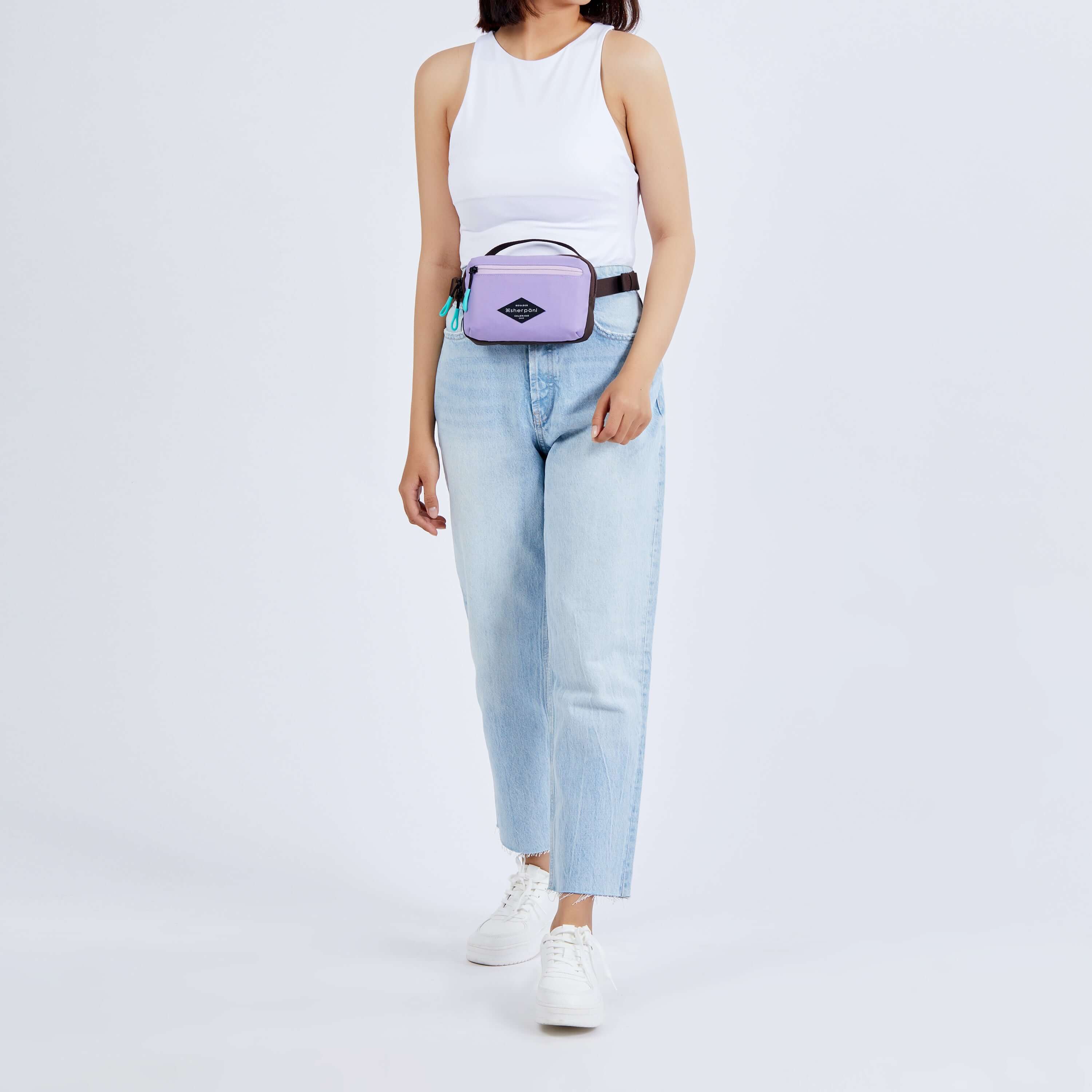 Close up view of a dark haired model. She is wearing a white tank top, jeans, white sneakers, and Sherpani's fanny pack, the Hyk in Lavender, as a fanny pack. 