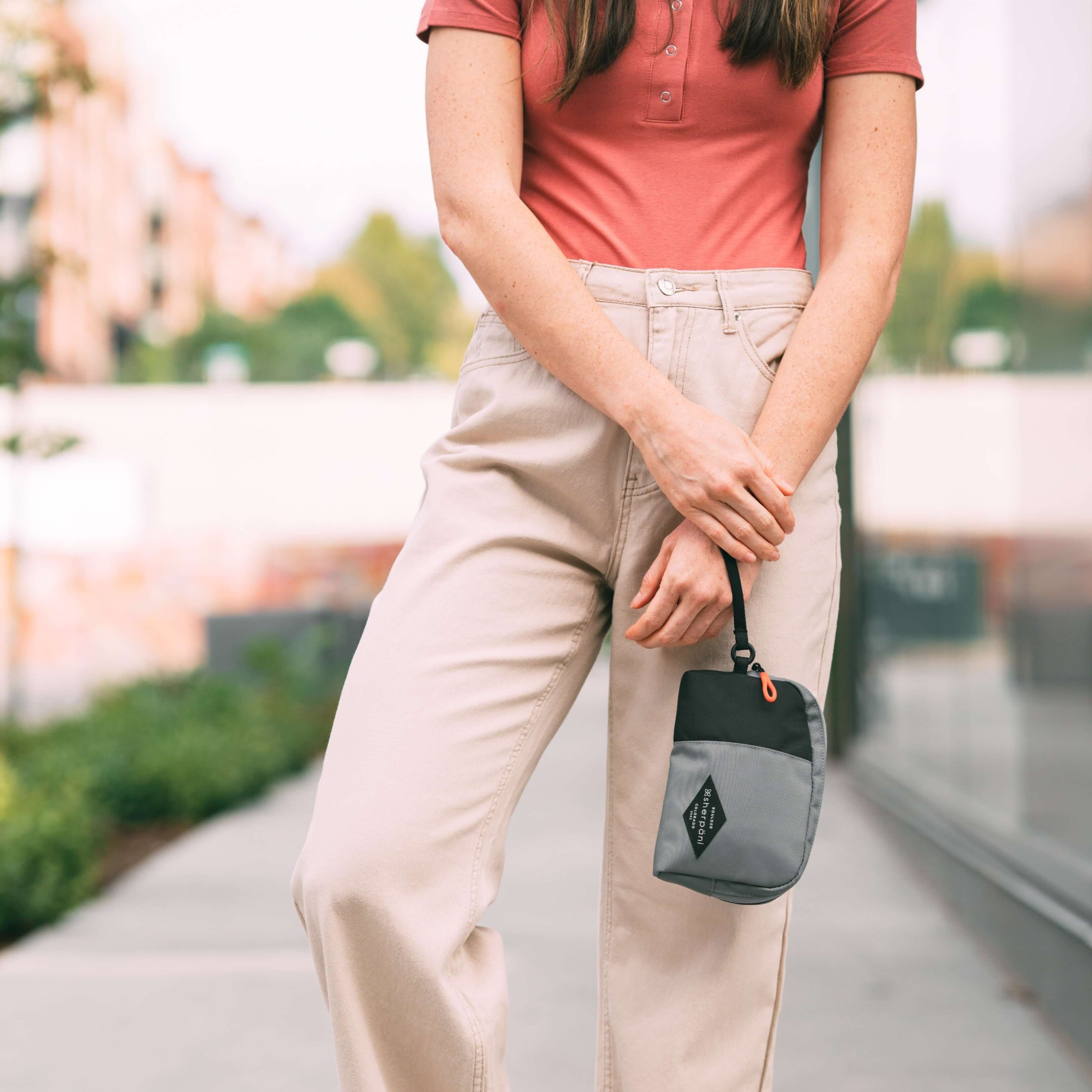 A brown haired woman stands outside on a path. She is wearing a salmon-colored top and tan pants. She carries Sherpani travel accessory, the Jolie in Stone, as a wristlet. 