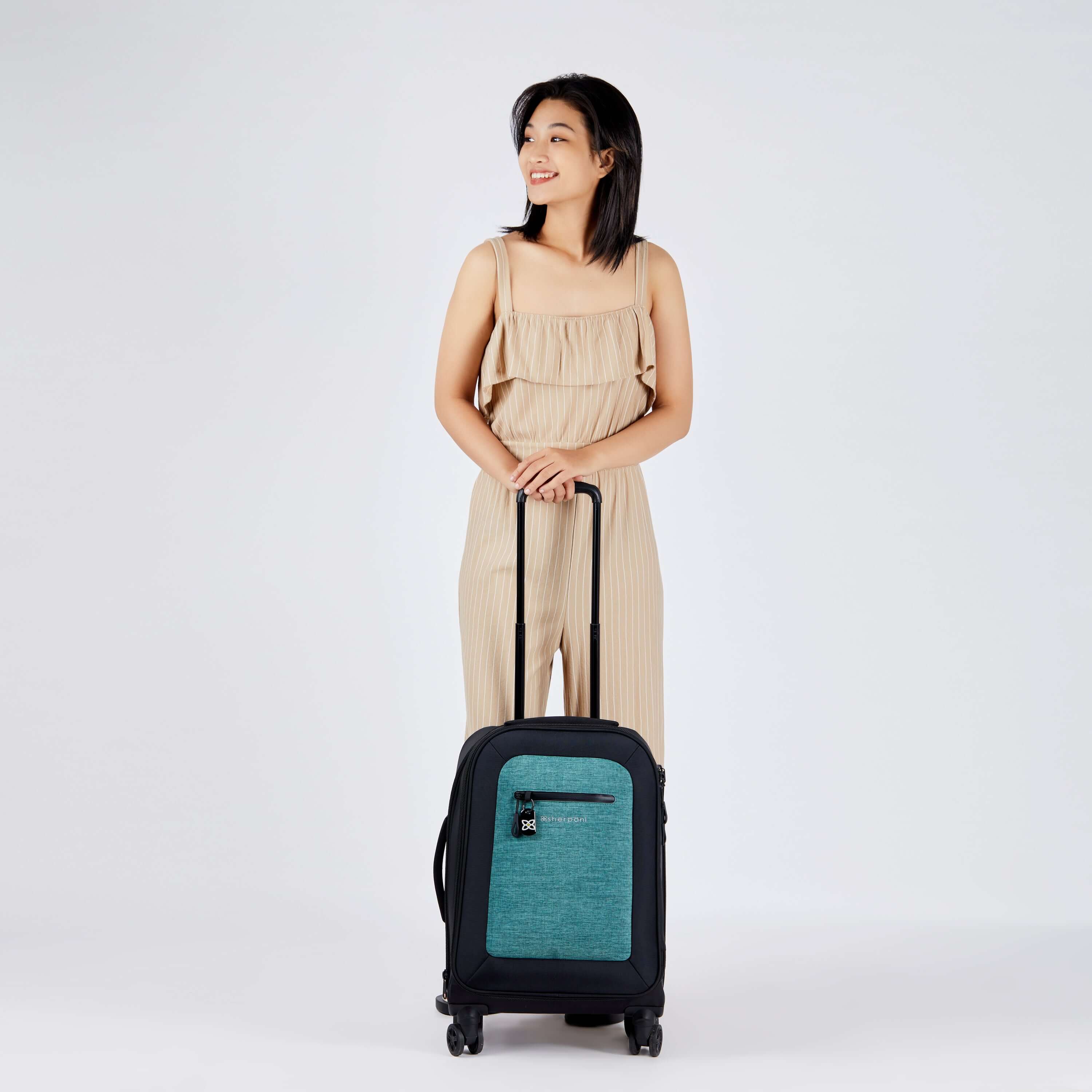 Full bodied view of dark haired model facing the camera and smiling over her right shoulder. She is wearing a tan jumpsuit. In front of her stands Sherpani's Anti-Theft luggage, the Latitude in Teal, which she is holding by the handle. 