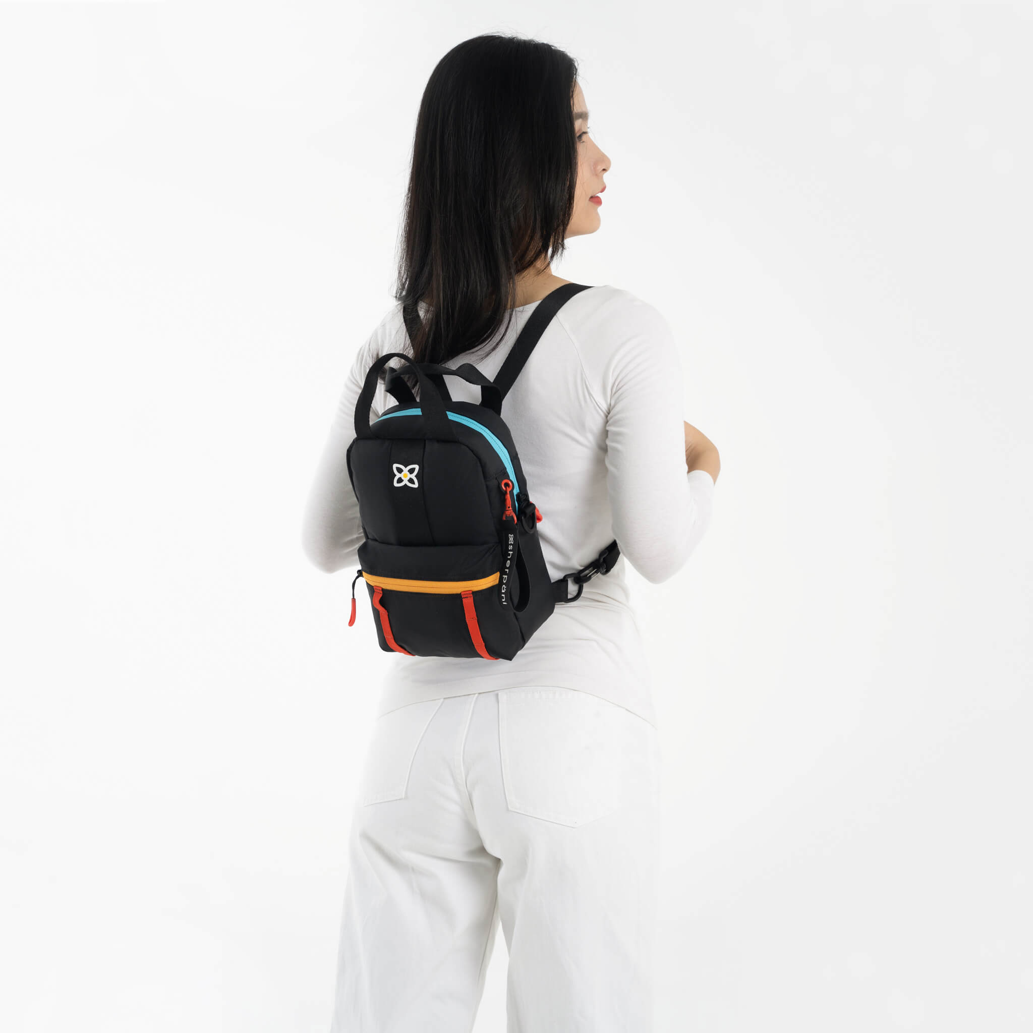A model facing to the back. She is wearing a white top, white pants and the Logan mini backpack in Chromatic. 