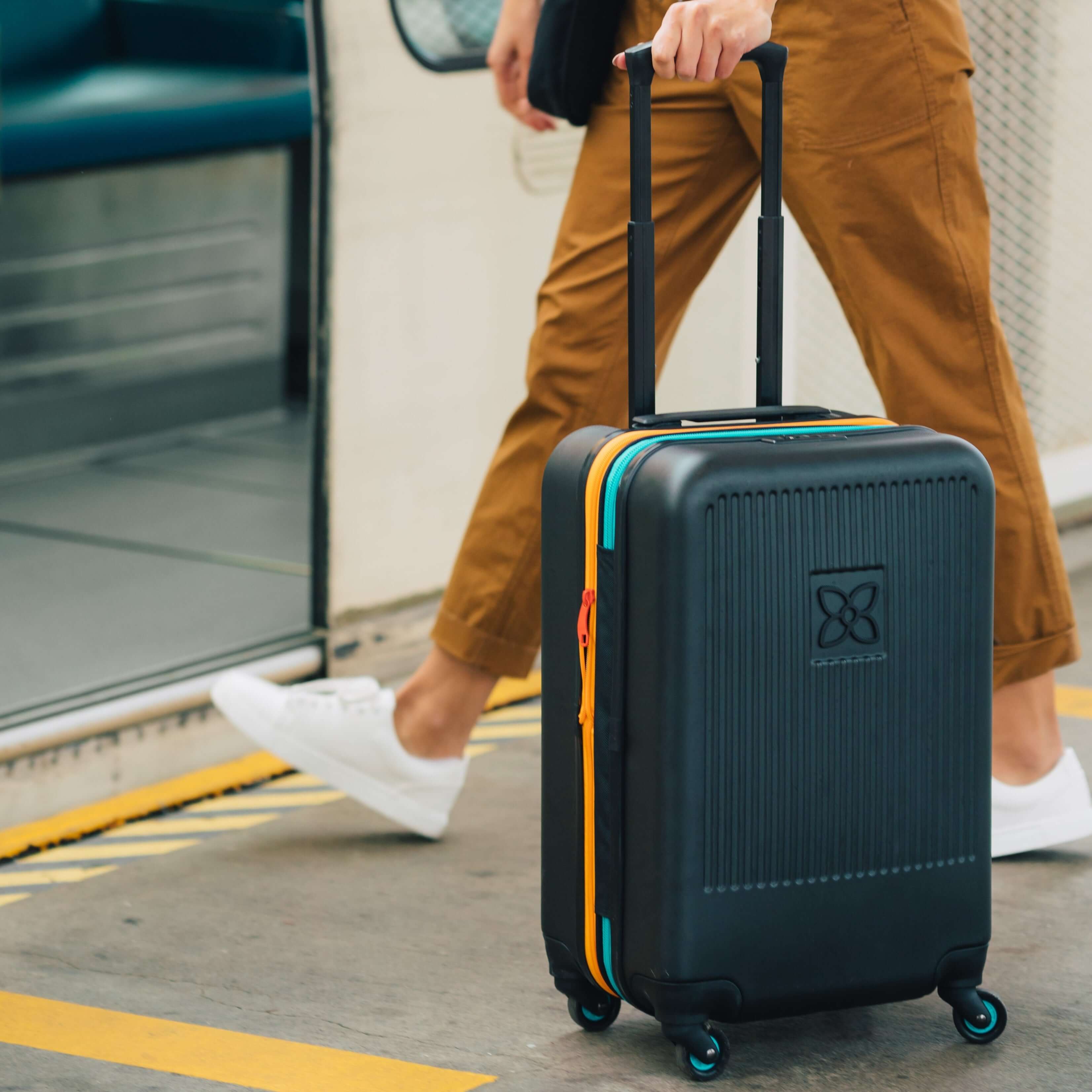 A woman stepping onto a train with Sherpani hard-shell carry-on luggage, the Meridian in Chromatic, in tow. 