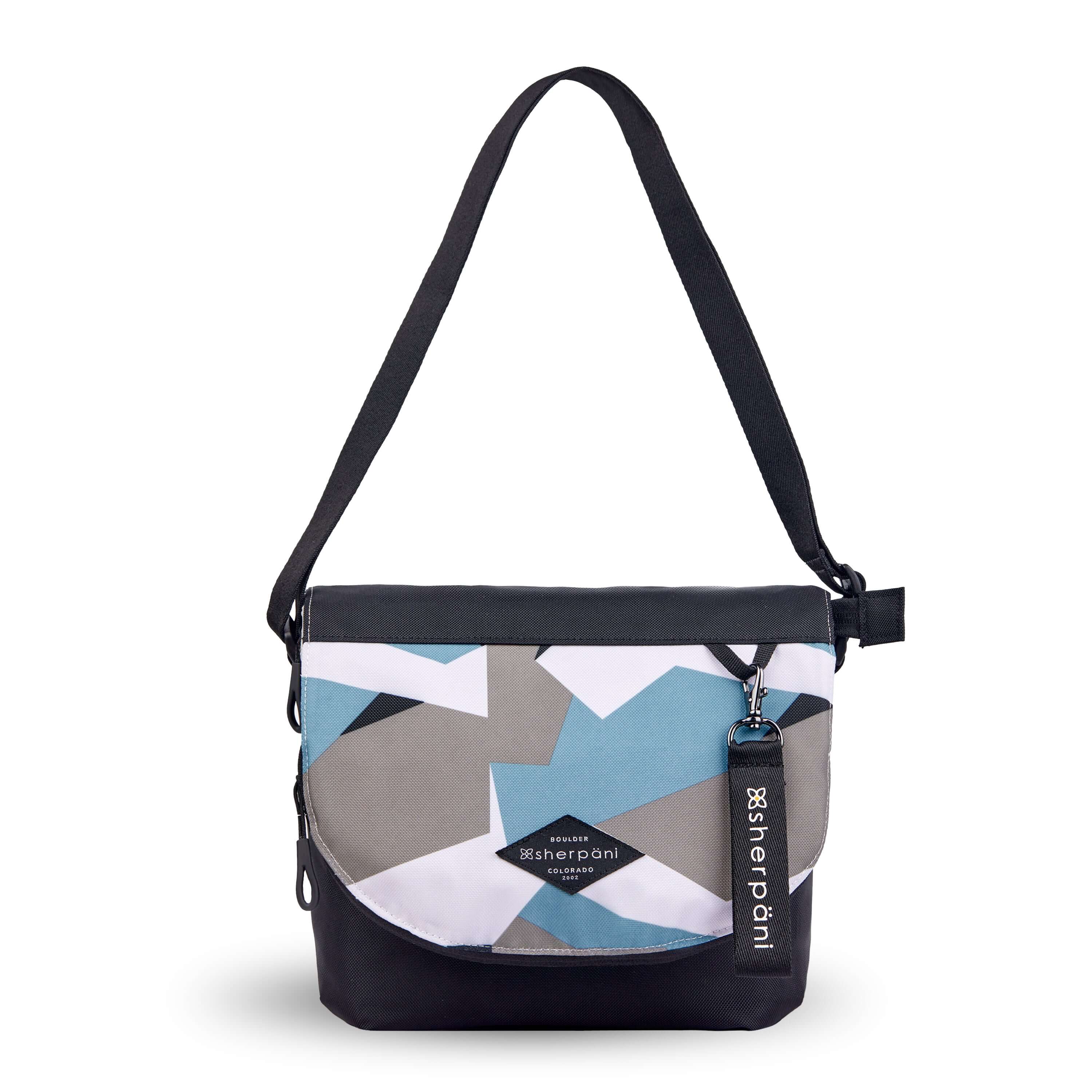 Flat front view of Sherpani crossbody, the Milli in Summer Camo. The messenger style bag is two toned: the layover flap is a camouflage pattern of light blue, gray and white, the remainder of the bag is black. Easy pull zippers are accented in black. The bag has an adjustable crossbody strap. A branded Sherpani keychain is clipped to a fabric loop in the upper right corner. 
