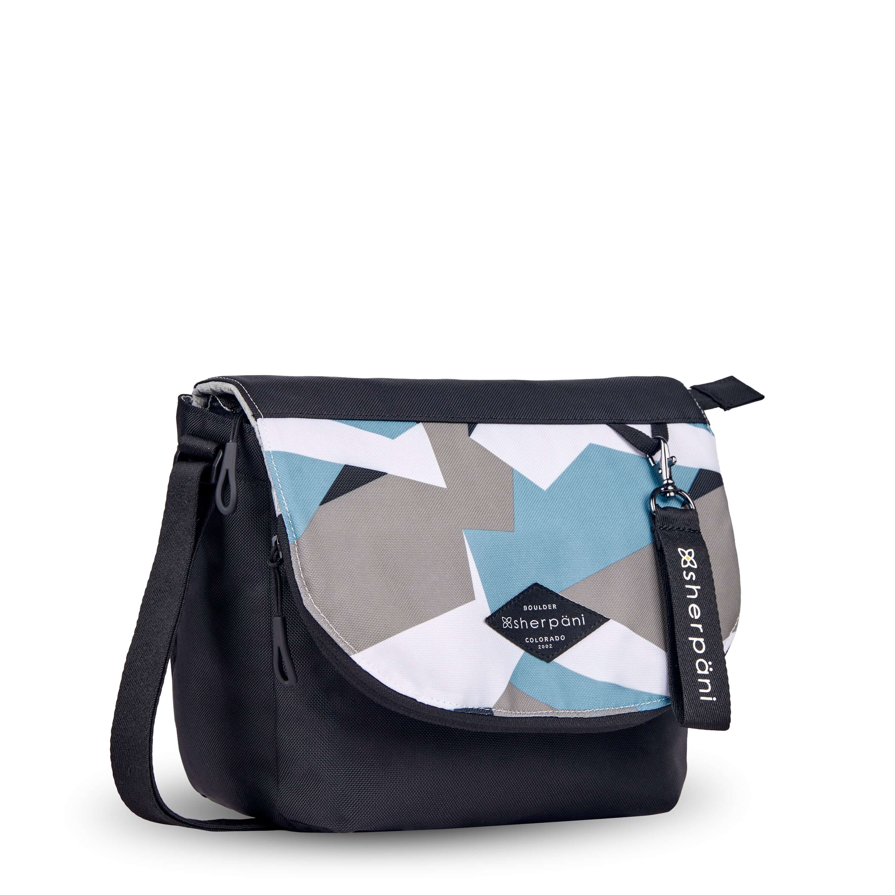 Angled front view of Sherpani crossbody, the Milli in Summer Camo. The messenger style bag is two toned: the layover flap is a camouflage pattern of light blue, gray and white, the remainder of the bag is black. Easy pull zippers are accented in black. The bag has an adjustable crossbody strap. A branded Sherpani keychain is clipped to a fabric loop in the upper right corner. 