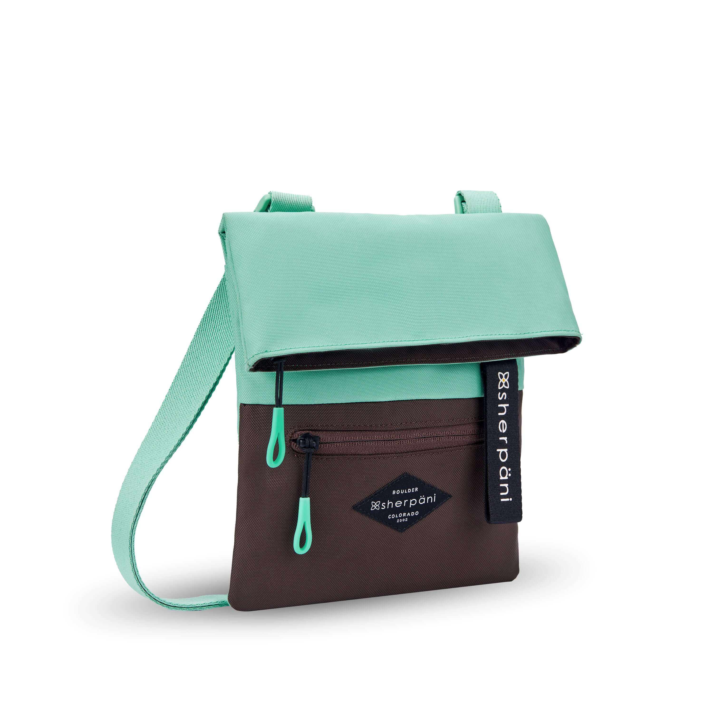Angled front view of Sherpani crossbody, the Pica in Seagreen. The bag is two toned: the top half is light green and the bottom half is brown. There is an external zipper pocket on the front panel. Easy-pull zippers are accented in light green. The top of the bag folds over creating a signature look. A branded Sherpani keychain is clipped to the upper right corner. The bag has an adjustable crossbody strap. #color_seagreen