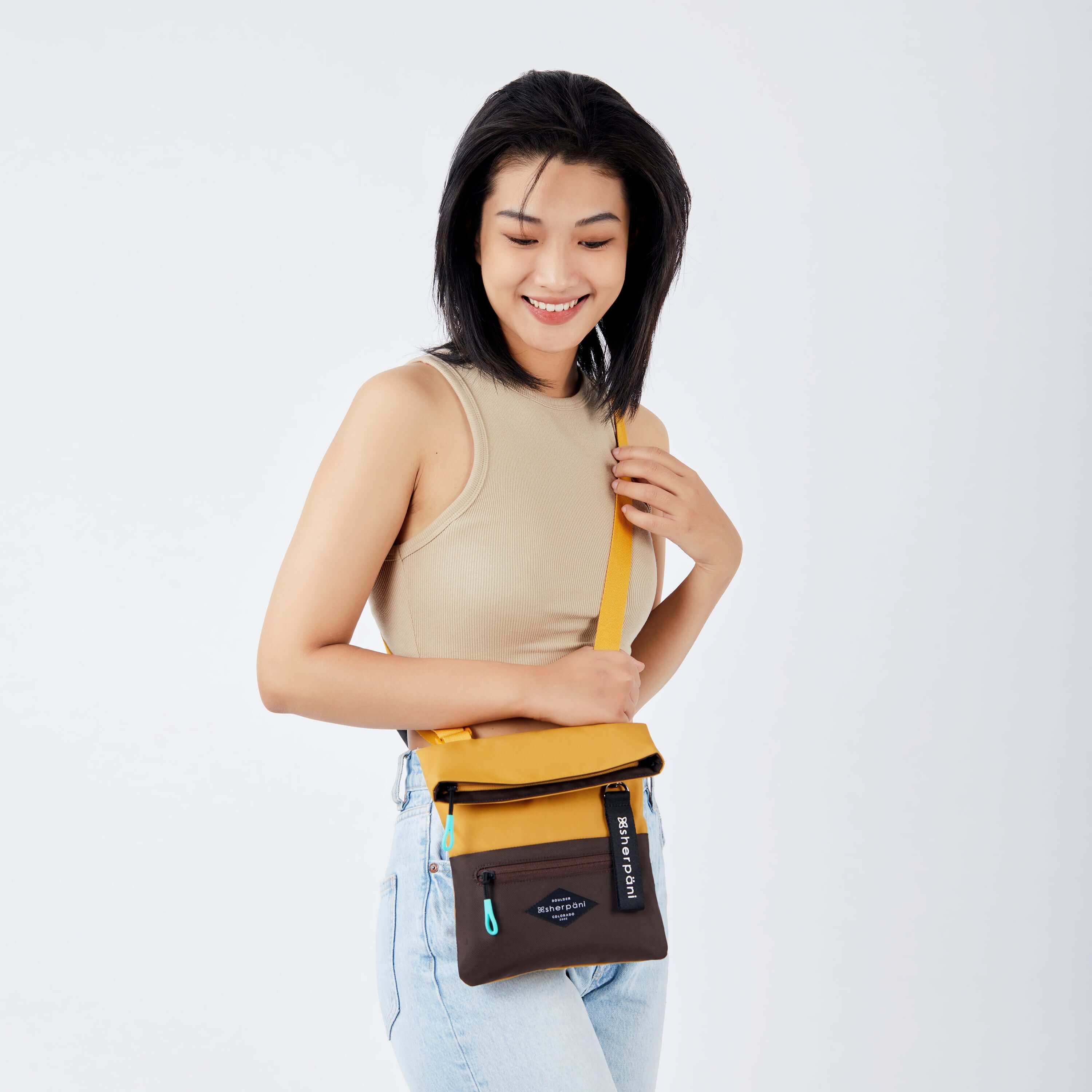 Close up view of a dark haired model facing the camera and smiling downward. She is wearing a tan crop top and jeans. She carries Sherpani crossbody, the Pica in Sundial, as a crossbody. #color_sundial v1