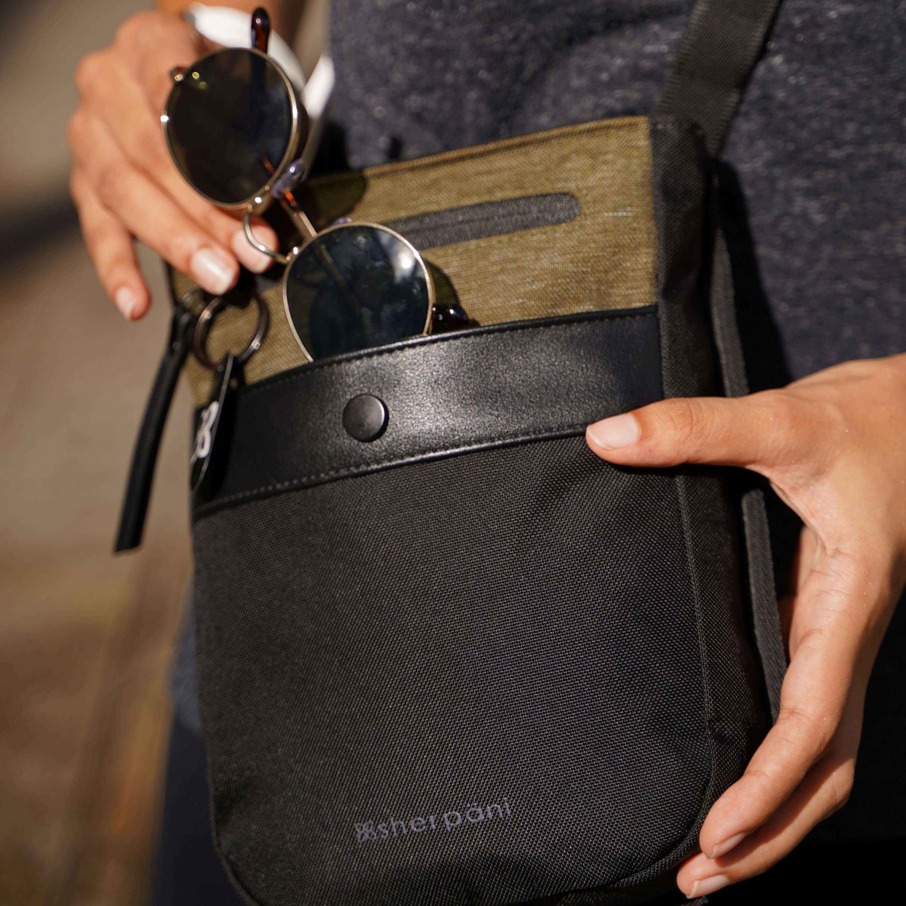 Close up view of a model's hands putting sunglasses into the front pocket of Sherpani's Anti-Theft bag the Prima AT in Loden. 