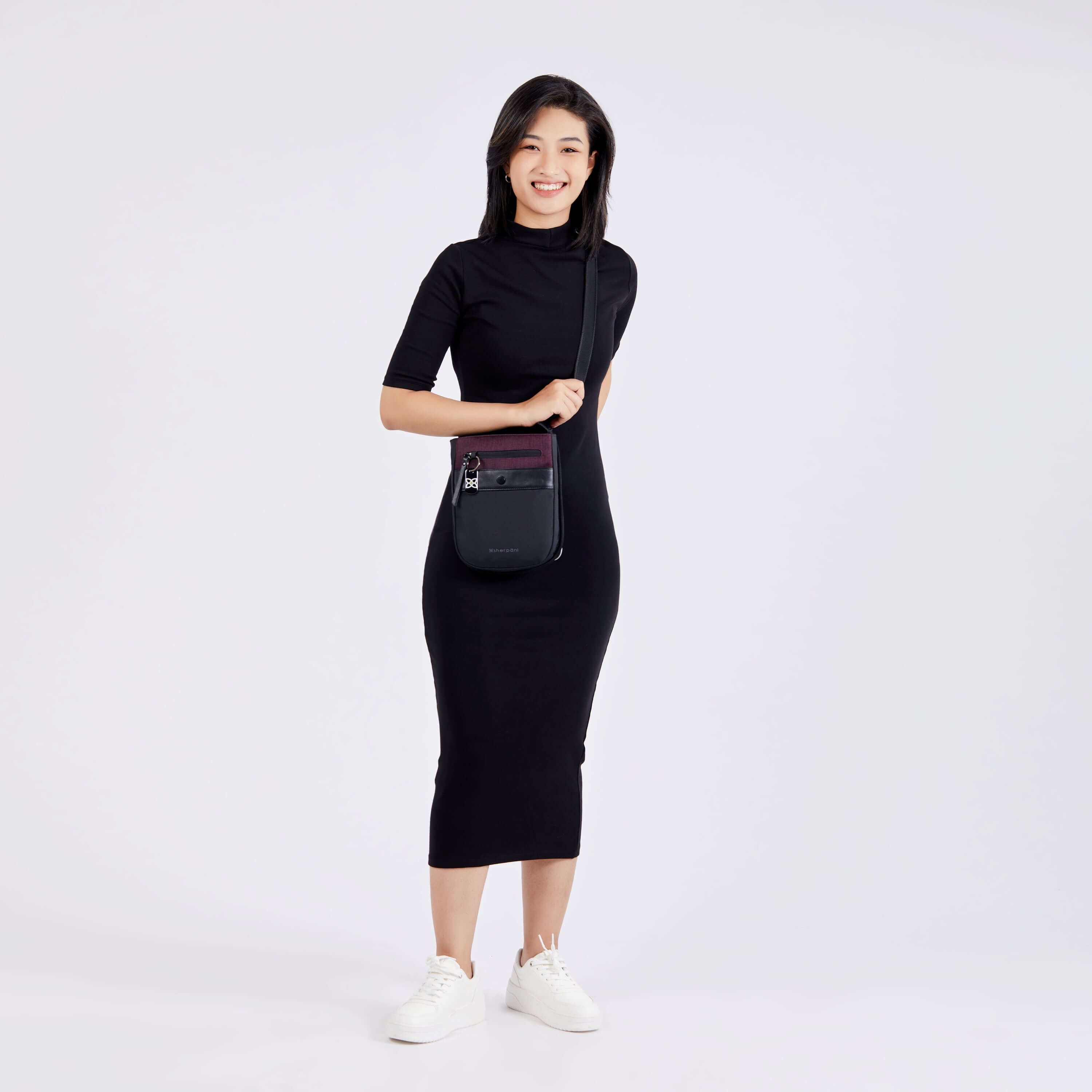 Full body view of a model facing the camera and smiling. She is wearing a black dress, white shoes and Sherpani's Anti-Theft crossbody the Prima AT in Merlot. 