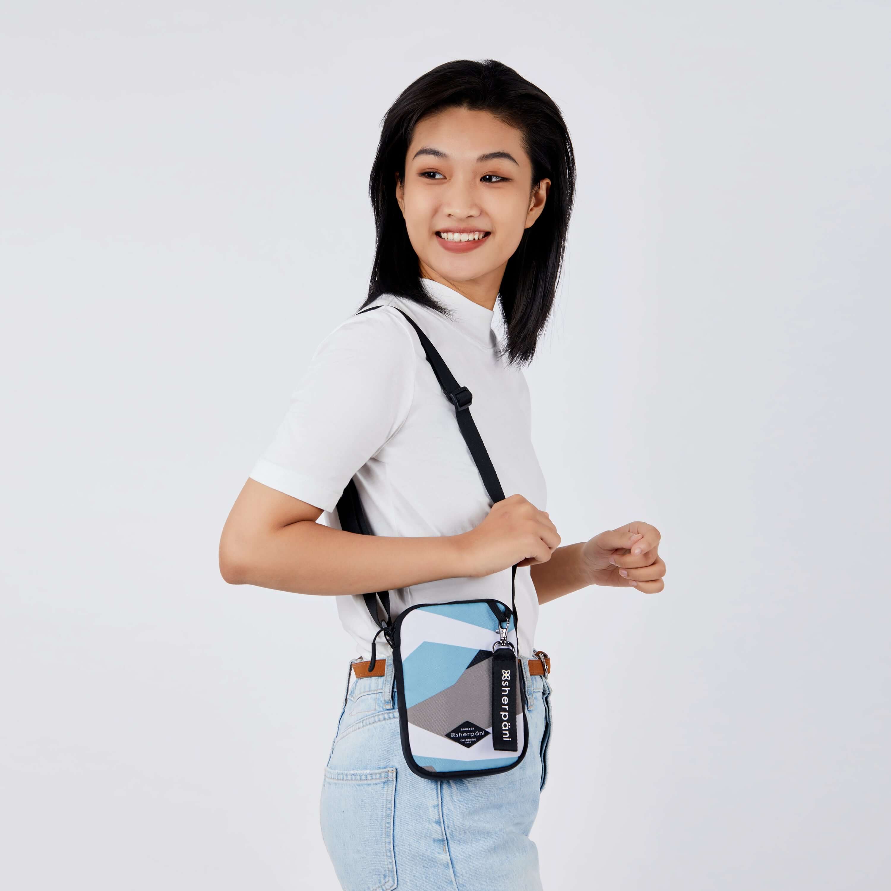 Close up view of a dark haired model facing the side and smiling over her right shoulder. She is wearing a white tee shirt and jeans. She carries Sherpani crossbody, the Rogue in Summer Camo, over her shoulder. 