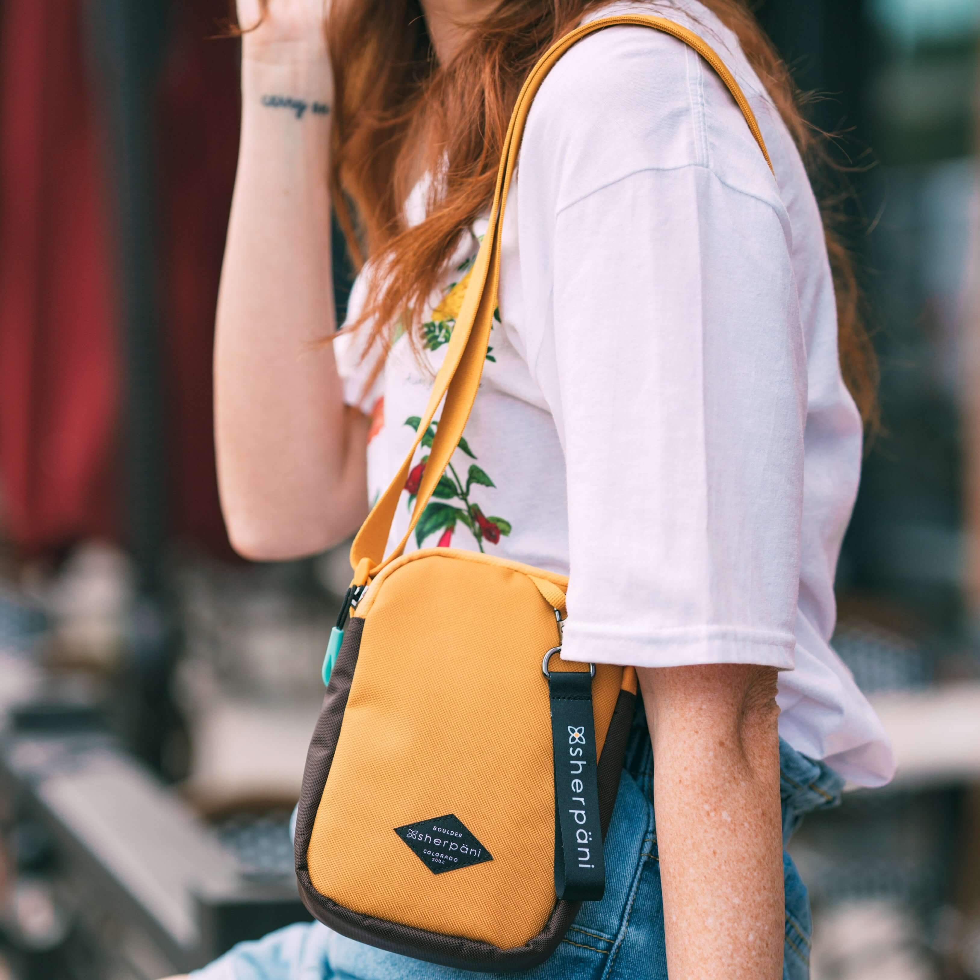 Close up view of a red haired woman outside. She is wearing a white shirt with flowers and jeans. She carries Sherpani crossbody, the Rogue in Sundial, over her shoulder. #color_sundial