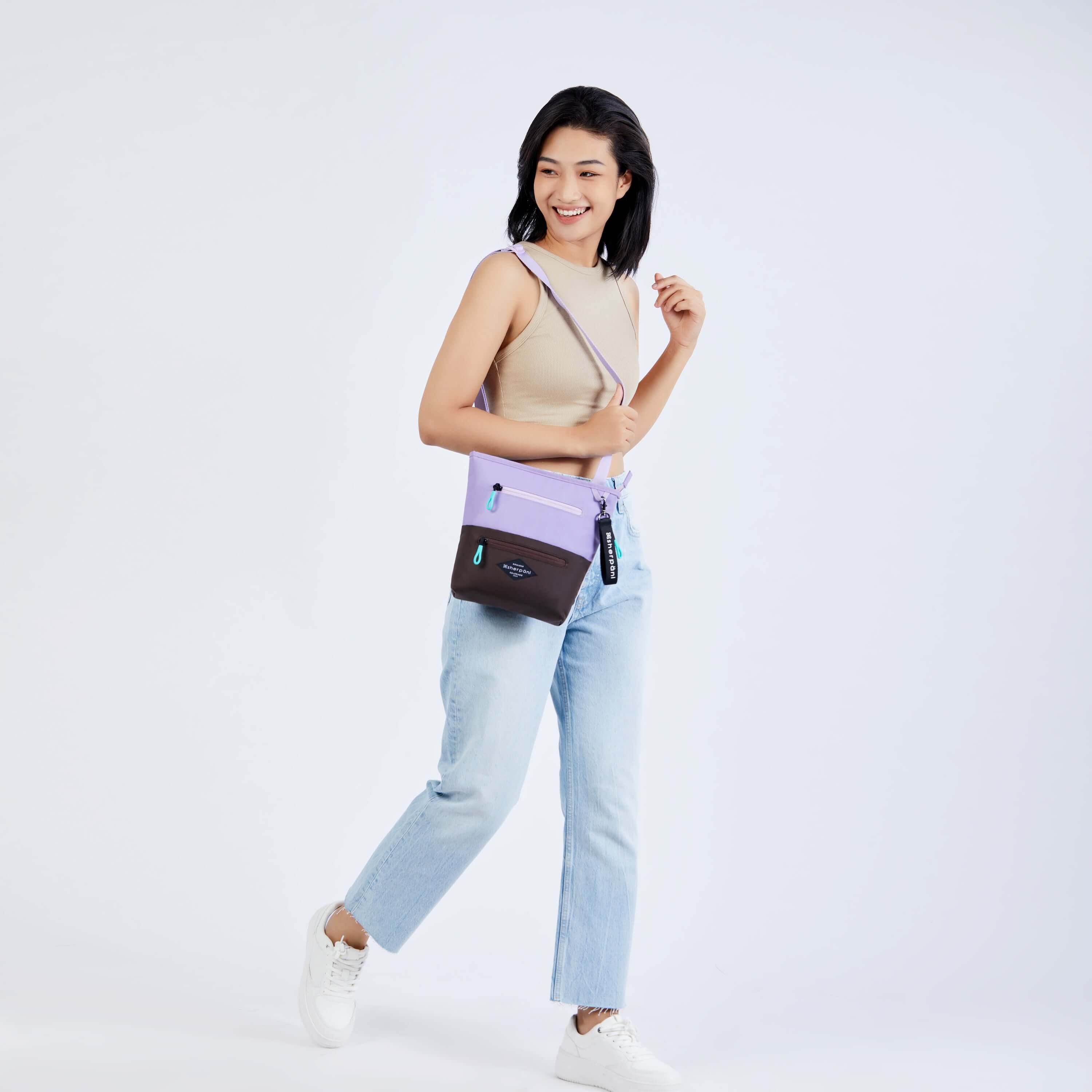 Full body view of a model walking to the side and smiling over her right shoulder. She is wearing a tan tank top, faded jeans and white sneakers. She carries Sherpani's crossbody, the Sadie in Lavender, over her shoulder. 
