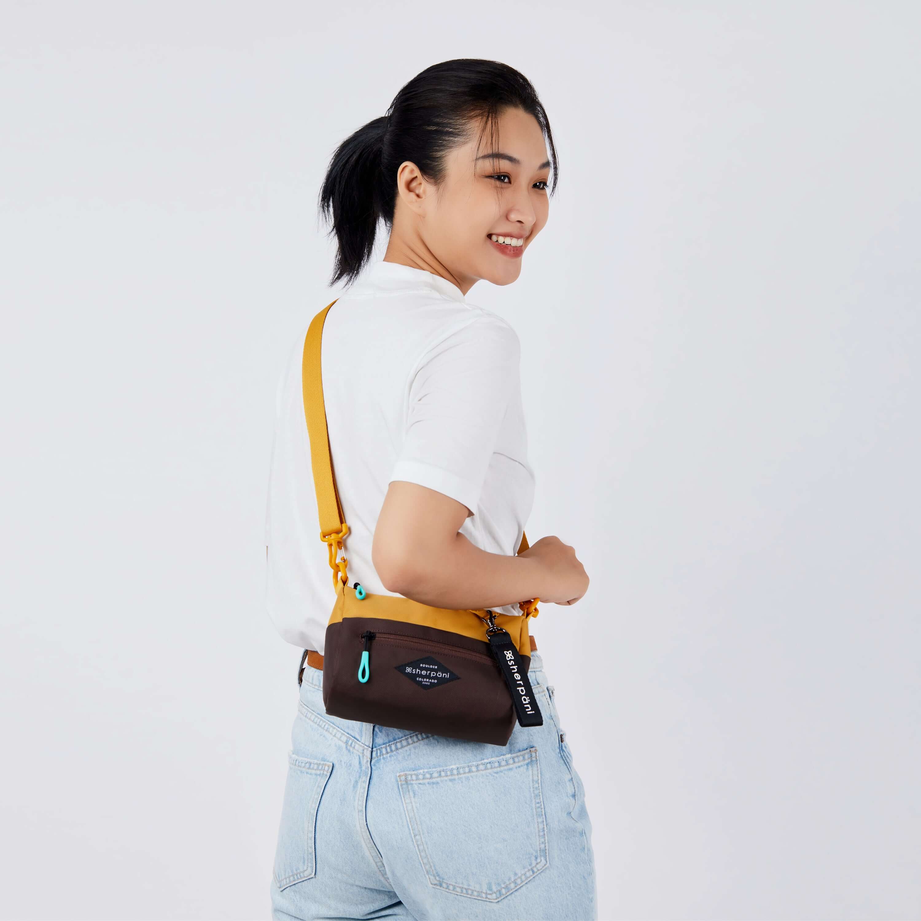 Close up view of a dark haired model facing away from the camera and smiling over her right shoulder. She is wearing a white tee shirt and jeans. She carries Sherpani's purse, the Skye in Sundial, as a crossbody. #color_sundial v1