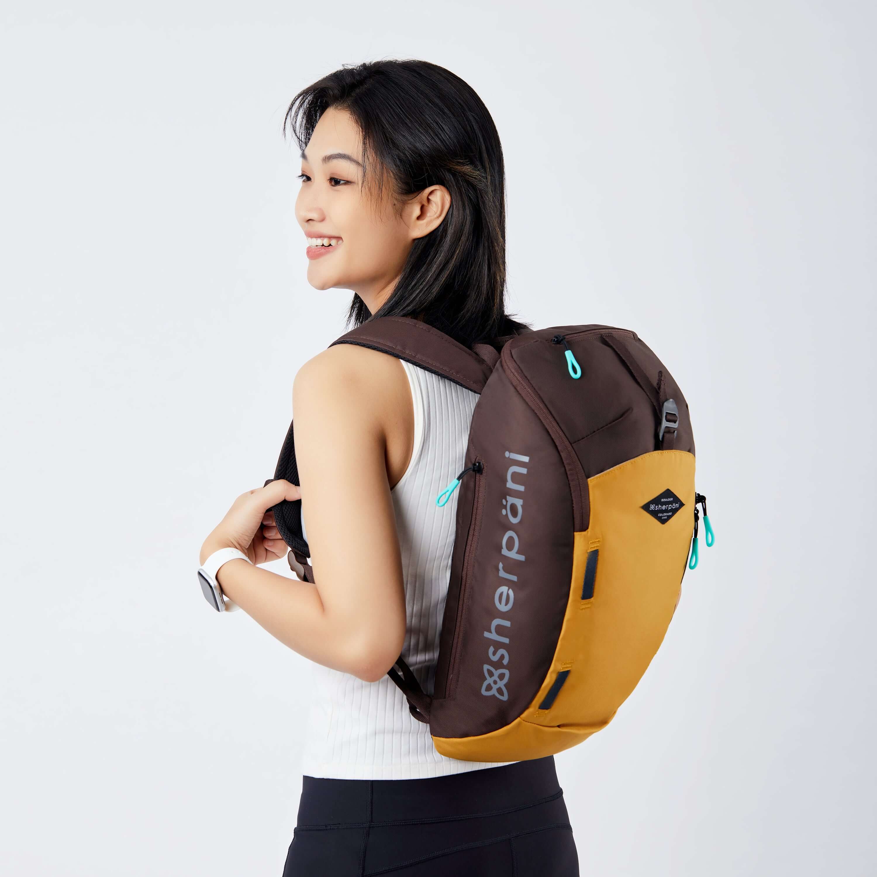 Close up view of a dark haired model facing away from the camera and smiling over her left shoulder. She is wearing a white tank top and black leggings. She carries Sherpani backpack, the Switch in Sundial, on her shoulders. 