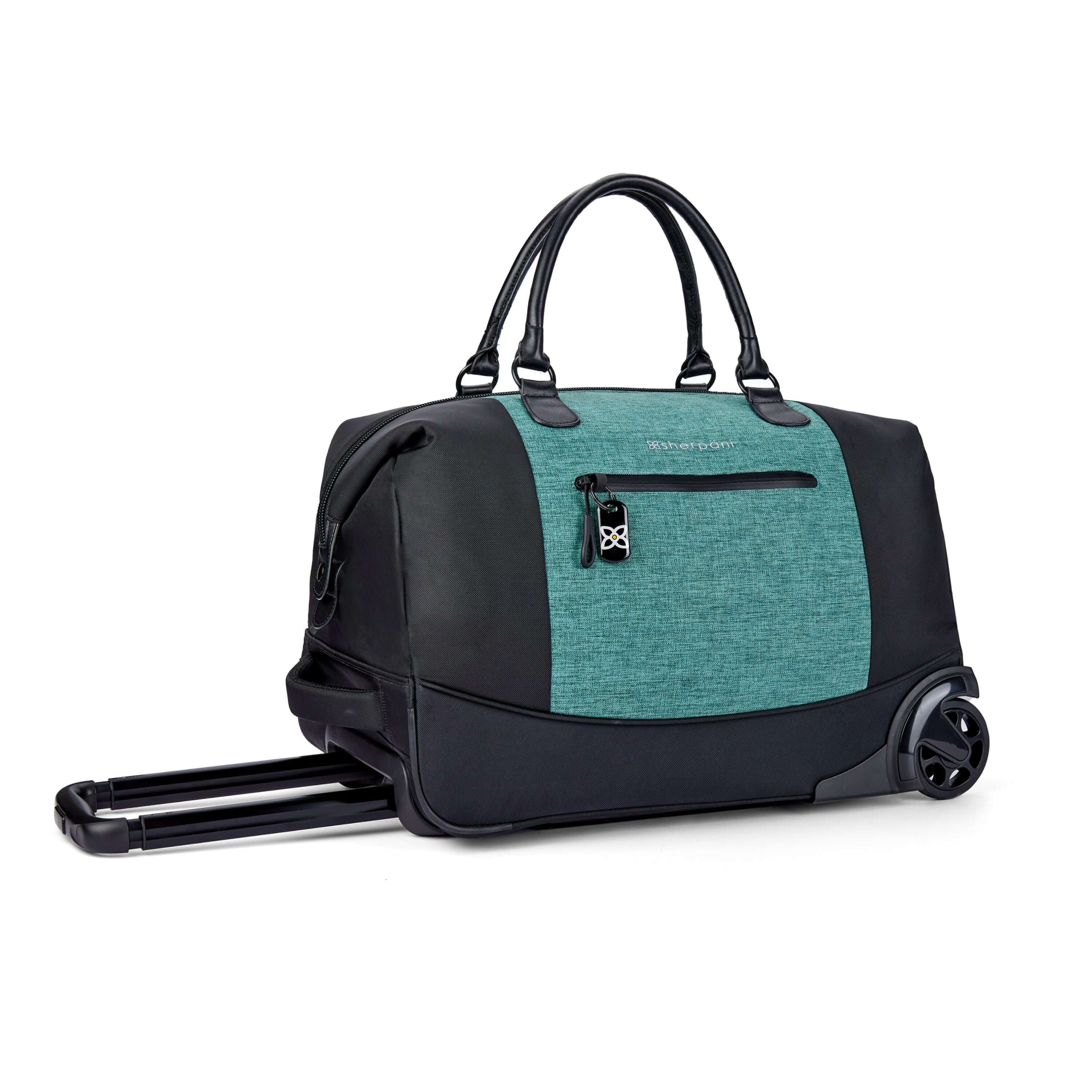 Angled front view of Sherpani’s Anti Theft rolling Duffle, the Trip in Teal, with vegan leather accents in black. The bag lies flat on the ground with the luggage handle extended on the left side and the rolling wheels shown on the right side. The bag features short tote handles at the top and an external zipper compartment on the front panel with locking zipper and ReturnMe tag. #color_teal