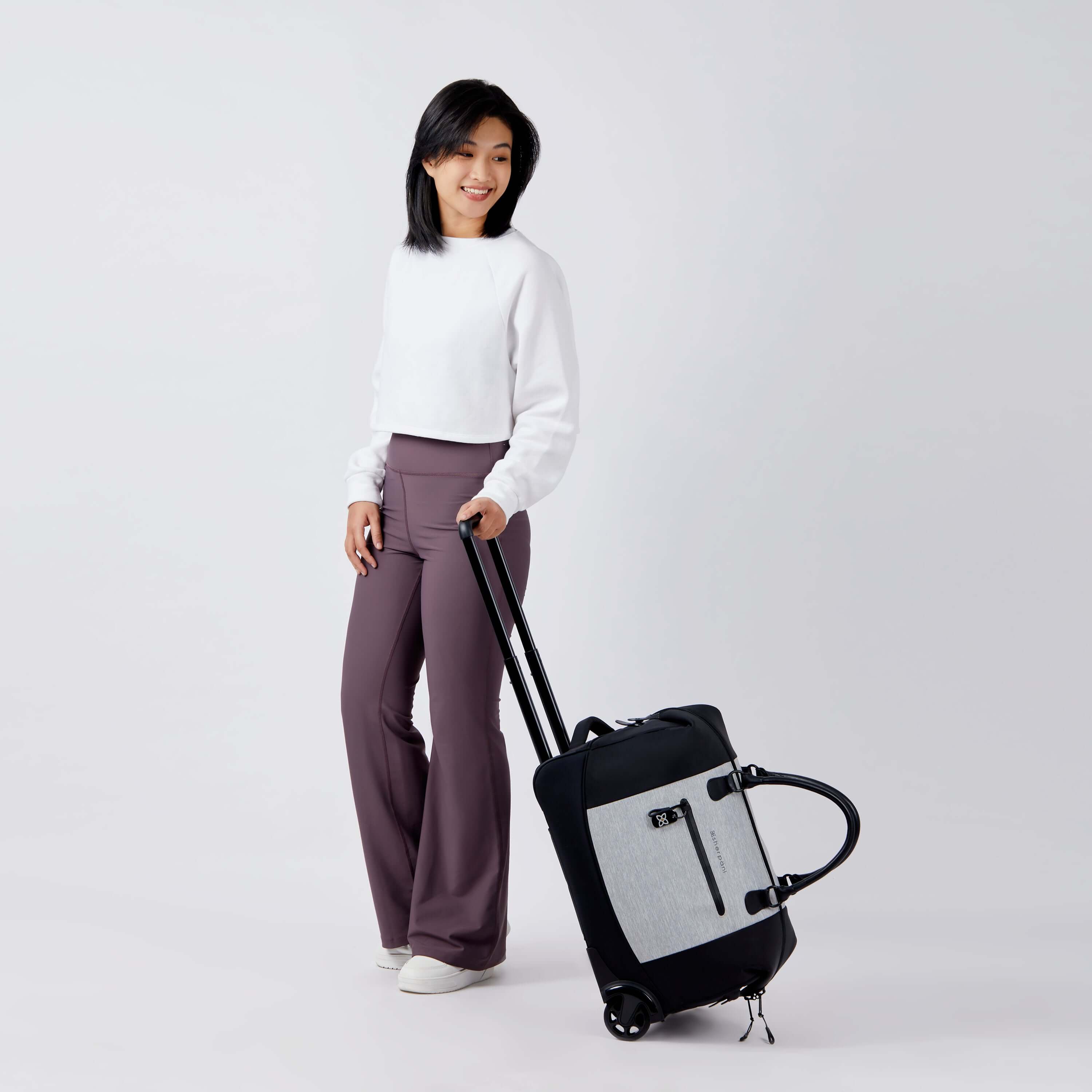 Full body view of a dark haired model facing the camera and smiling. She is wearing a white shirt and purple leggings. She is holding the luggage handle of Sherpani's Anti-Theft rolling duffle the Trip in Sterling. 