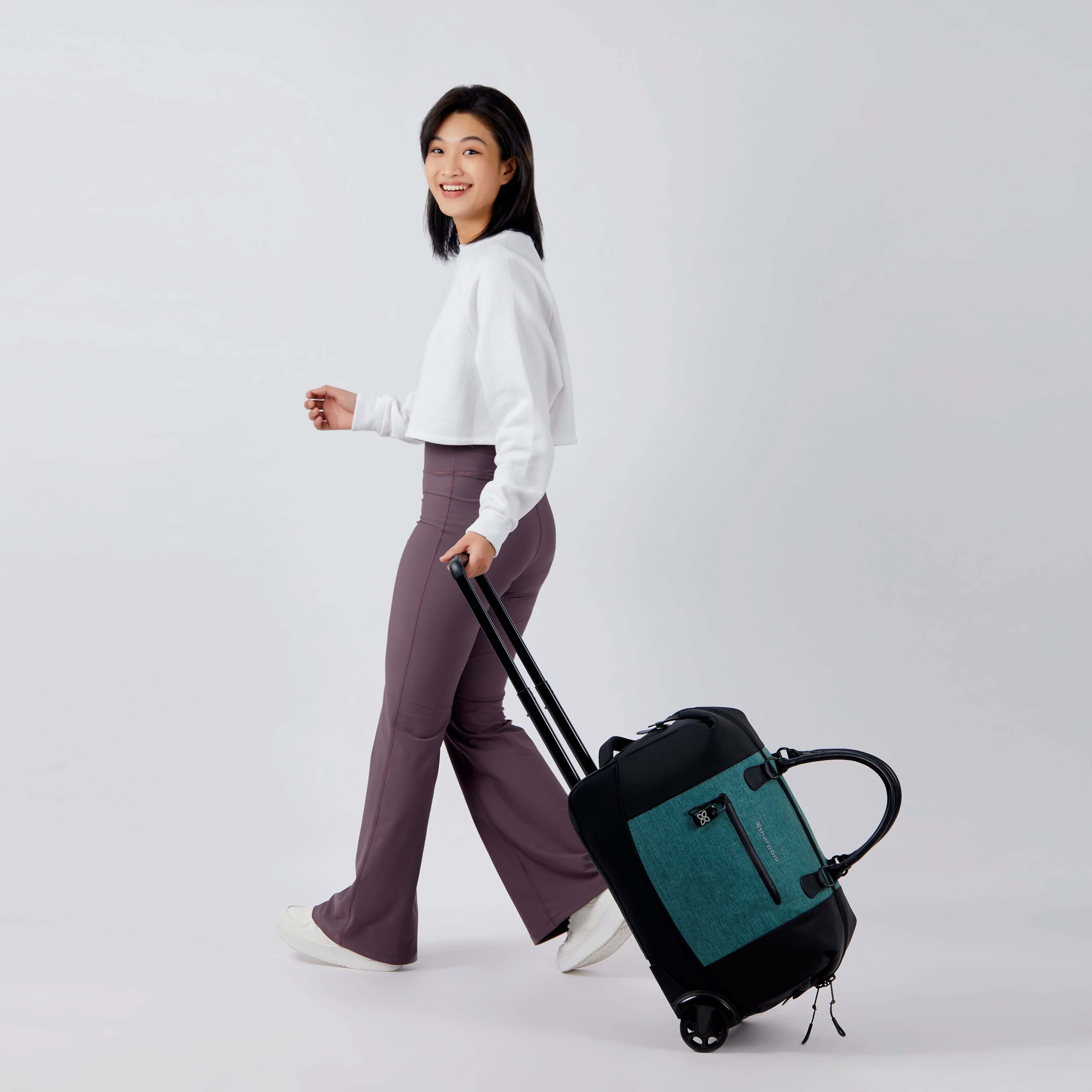 Full bodied view of a dark haired model facing the side and is smiling over her left shoulder at the camera. She is wearing a white shirt and purple leggings. She is holding the luggage handle of Sherpani's Anti-Theft rolling duffle the Trip in Teal as the bag rolls along beside her. #color_teal