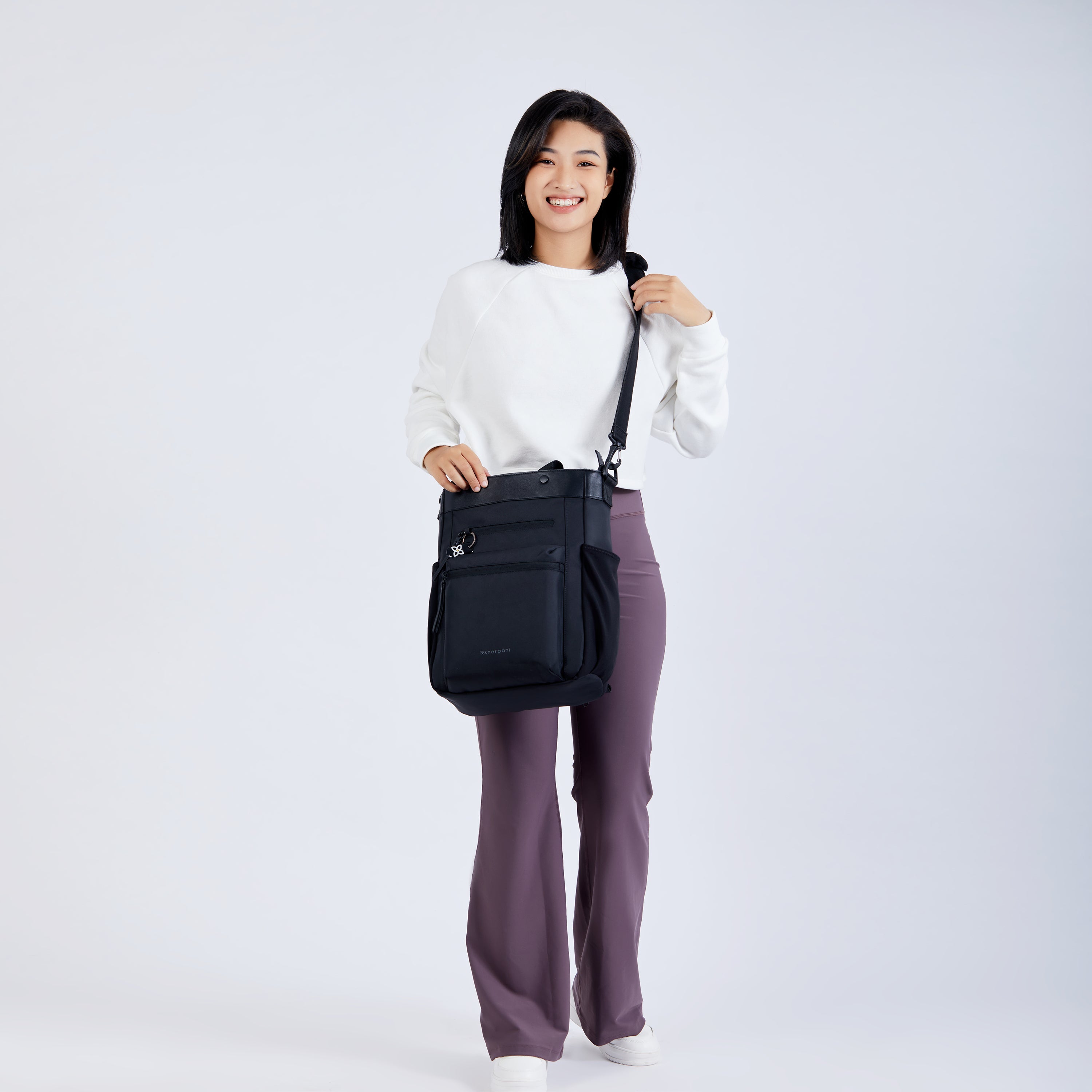 A dark haired model faces the camera and smiles. She is wearing a white shirt, purple leggings and white shoes. She carries Sherpani's Anti-Theft bag, the Soleil in Carbon, as a crossbody.