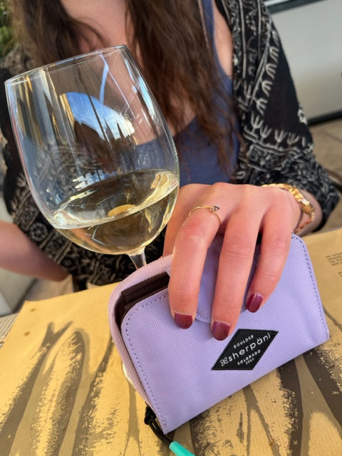 A woman sits at table outdoors. A glass of Vinho Verde white wine is on the table in front of her. She holds Sherpani wallet, the Barcelona in Lavender, in her hand next to the wine.