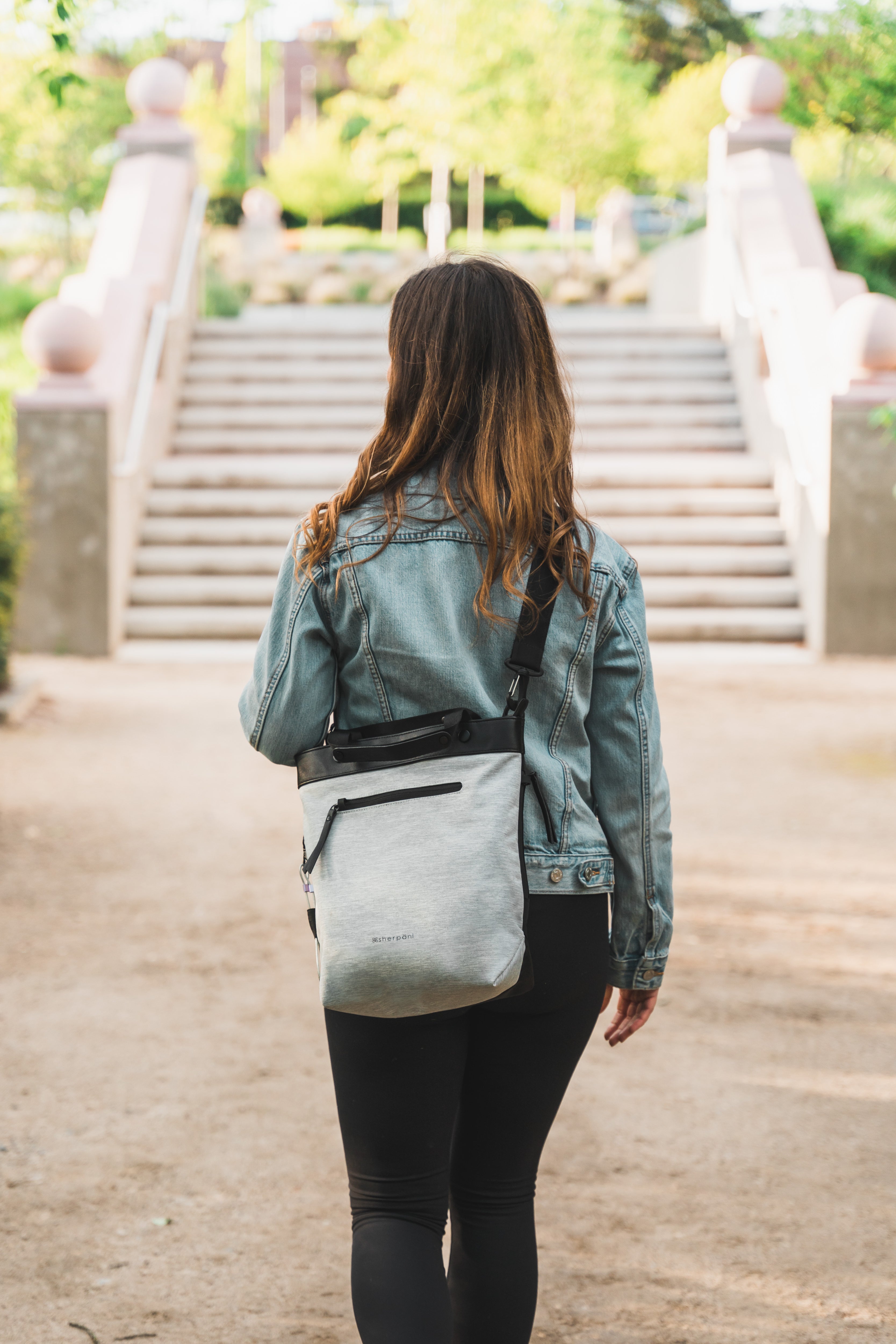 A brown-haired woman walks away from the camera toward an outdoor staircase. She is carrying Sherpani's Anti-Theft bag, the Geo, as a crossbody.