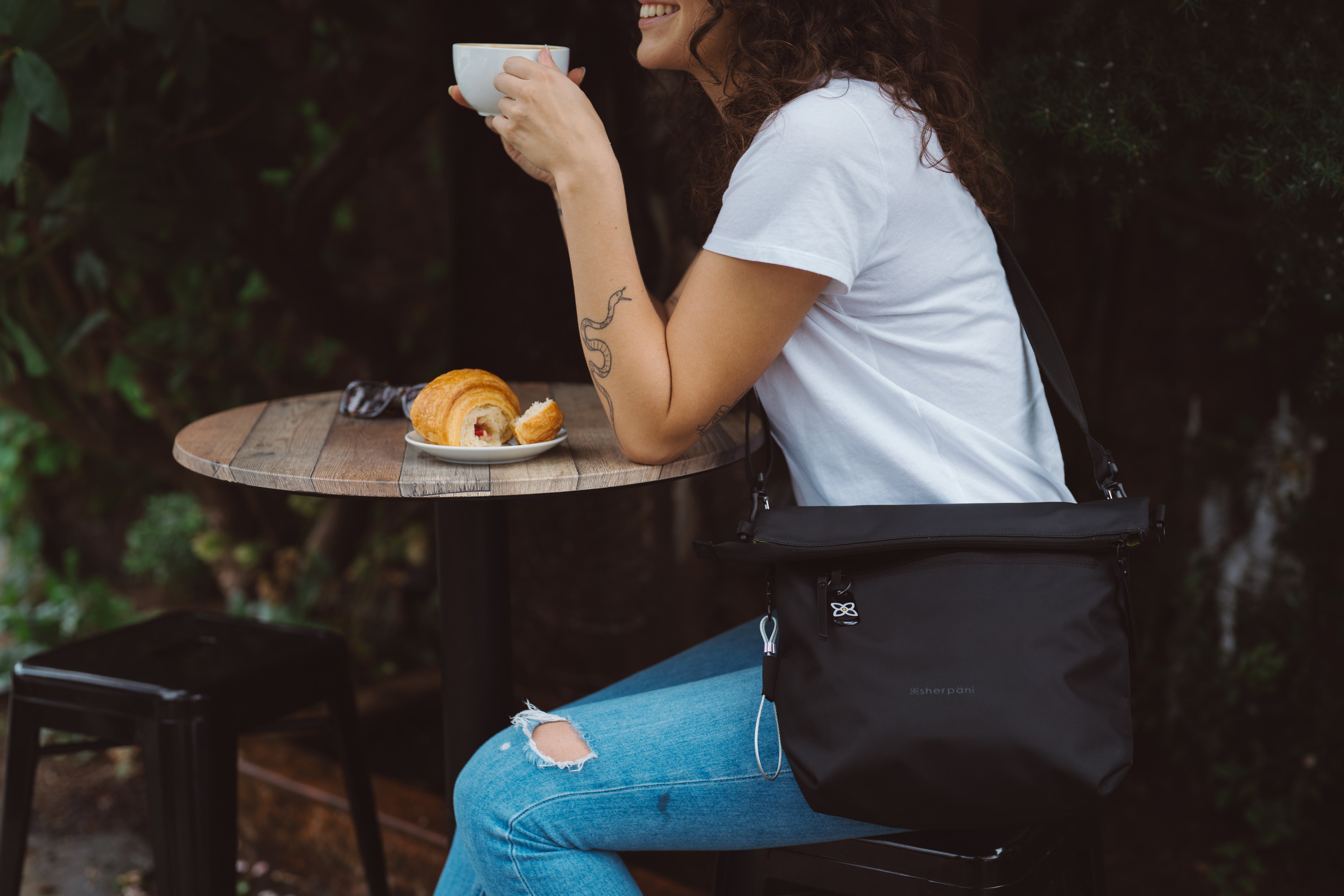 A woman sitting at an outdoor table drinking a coffee. She is wearing Sherpani crossbody Anti-Theft bag, the Vale.