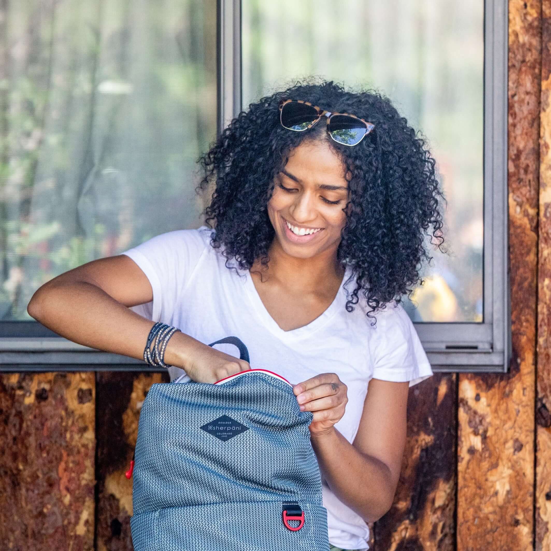 A curly haired woman sits in front of a wooden wall outside. She is wearing a white tee shirt and smiling downward. She is unzipping the main compartment of Sherpani mesh backpack, the Adaline.