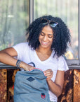 A curly haired woman sits in front of a wooden wall outside. She is wearing a white tee shirt and smiling downward. She is unzipping the main compartment of Sherpani mesh backpack, the Adaline.