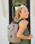 A blonde woman stands outside in front of a house and smiles over her right shoulder at the camera. She is wearing a green dress. She carries Sherpani mesh backpack, the Adaline.