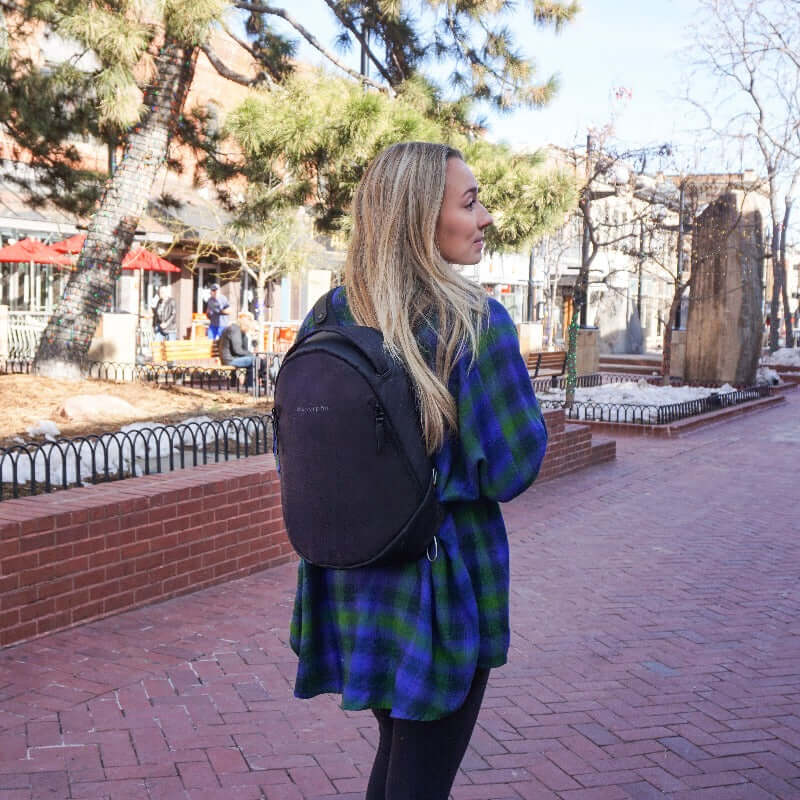 A blonde woman facing away from the camera walks down the street of an outdoor mall. She is wearing a blue and green flannel, black leggings and Sherpani&#39;s Anti-Theft backpack, the Presta in Merlot.