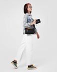 Close up view of a dark haired model facing the camera and looking off to the side. She is wearing a jean jacket and white pants. She carries Sherpani crossbody, the Zoom in Chromatic, as a crossbody. She holds Sherpani wallet, the Barcelona in Raven, in her right hand.